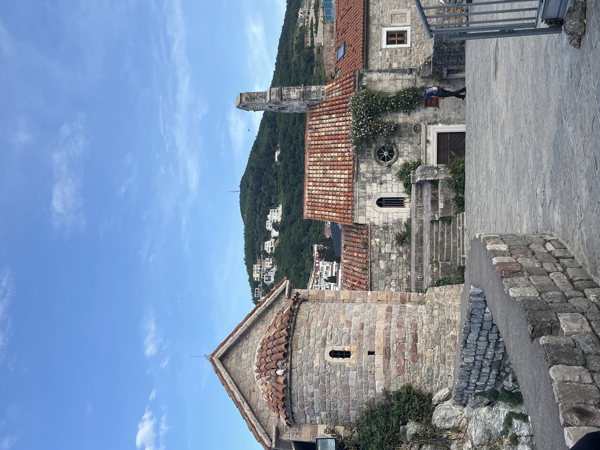 The image shows a view in Budva, Montenegro, focusing on the architecture of the old town. In the foreground is a stone building with a tiled roof. A taller structure with a bell tower is visible in the midground. The building is constructed of stone, with a red-tiled roof. Green vegetation grows on the walls of the building. The landscape features a tree-covered hill in the background. A small number of residential houses are visible in the midground. The sky is light blue with clouds. A man is walking near a stone building.