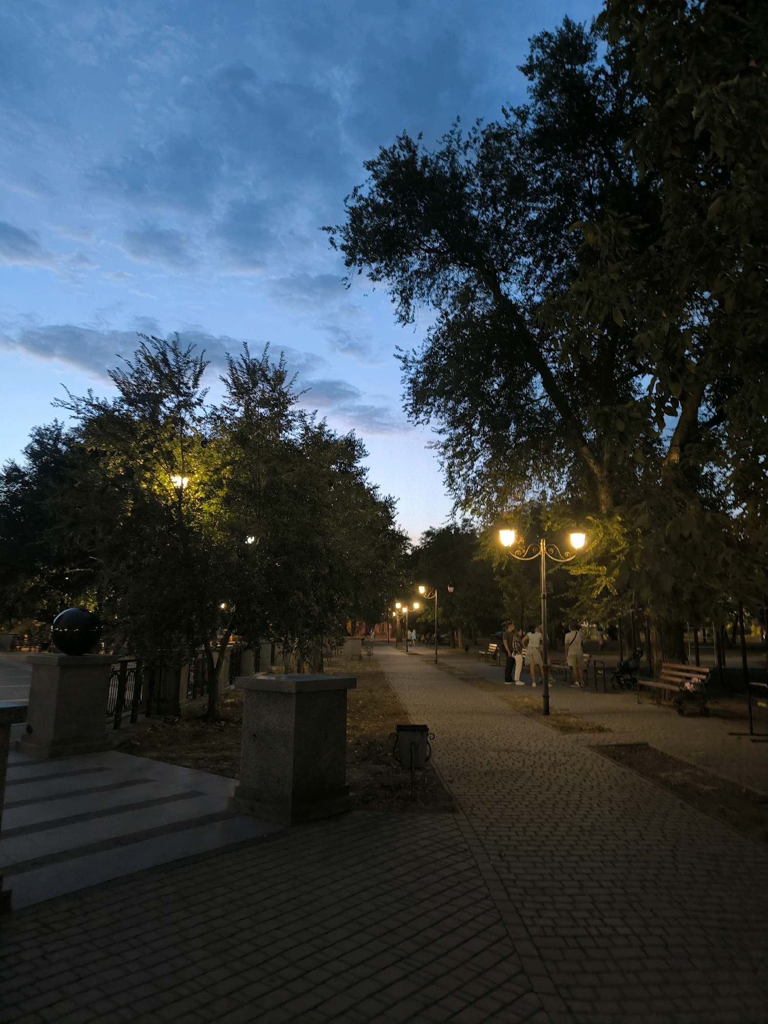 The image depicts a park pathway in Mykolaiv, Ukraine, at dusk. A paved walkway with cobblestone texture extends into the distance, lined by trees on either side. Several street lamps with ornate designs cast a warm yellow glow, illuminating portions of the path and the surrounding foliage. Some people are visible walking or standing on the pathway and on benches situated along the route. On the left side of the image, a wide stone staircase leads down to a lower level, with a large, dark spherical object on a pedestal. The sky above is a dusky blue with scattered clouds, indicating the transition from day to night.