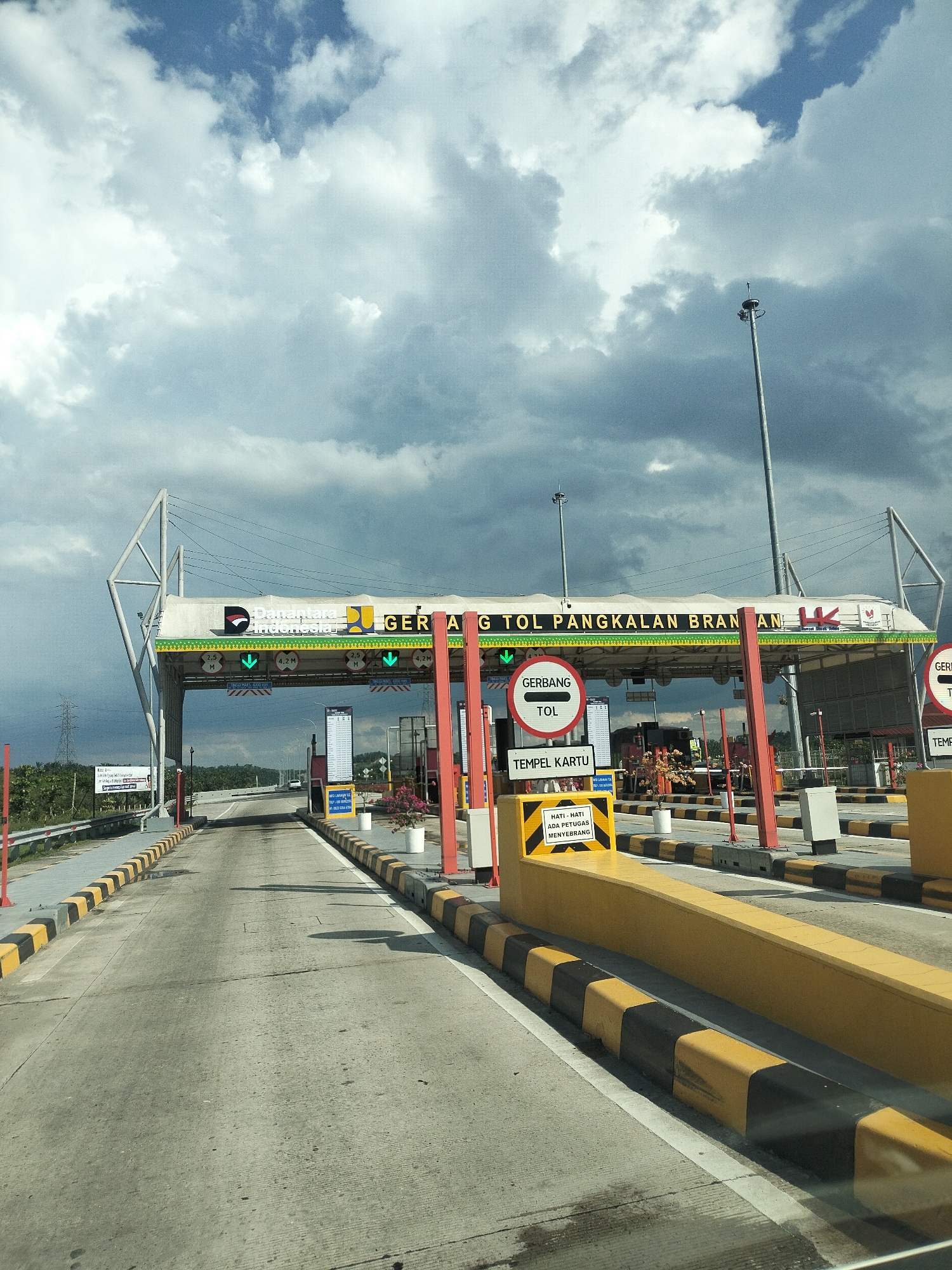 The image depicts a toll gate on a highway in Tangkahan Durian, Indonesia. The toll gate has several lanes, marked with yellow and black stripes, leading towards the booths. Overhead, a large structure displays "GERBANG TOL PANGKALAN BRAN AN". Red poles support the toll gate structure. Green arrows indicate open lanes, while signs like "GERBANG TOL" and "TEMPEL KARTU" are visible. Yellow and black barricades line the road. In the background, trees and some buildings are visible under a cloudy sky. There are no people clearly visible in the image.