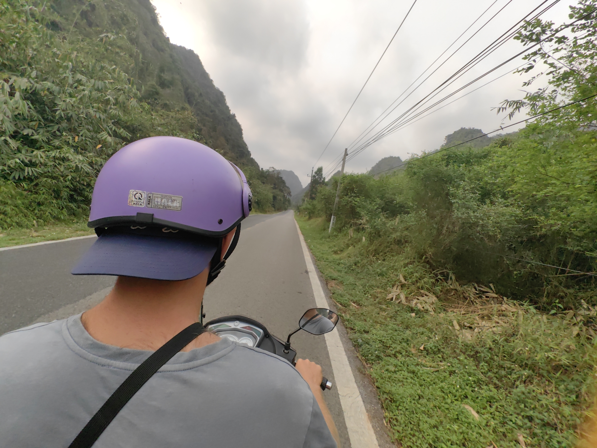 A person wearing a lavender helmet and grey t-shirt is riding a scooter on a paved road. The road is flanked by lush green vegetation and trees on both sides, with mountains visible in the background under a cloudy sky. Utility poles with overhead wires run along the right side of the road. The location appears to be in a rural or mountainous area, potentially near Hải Phòng, Vietnam.