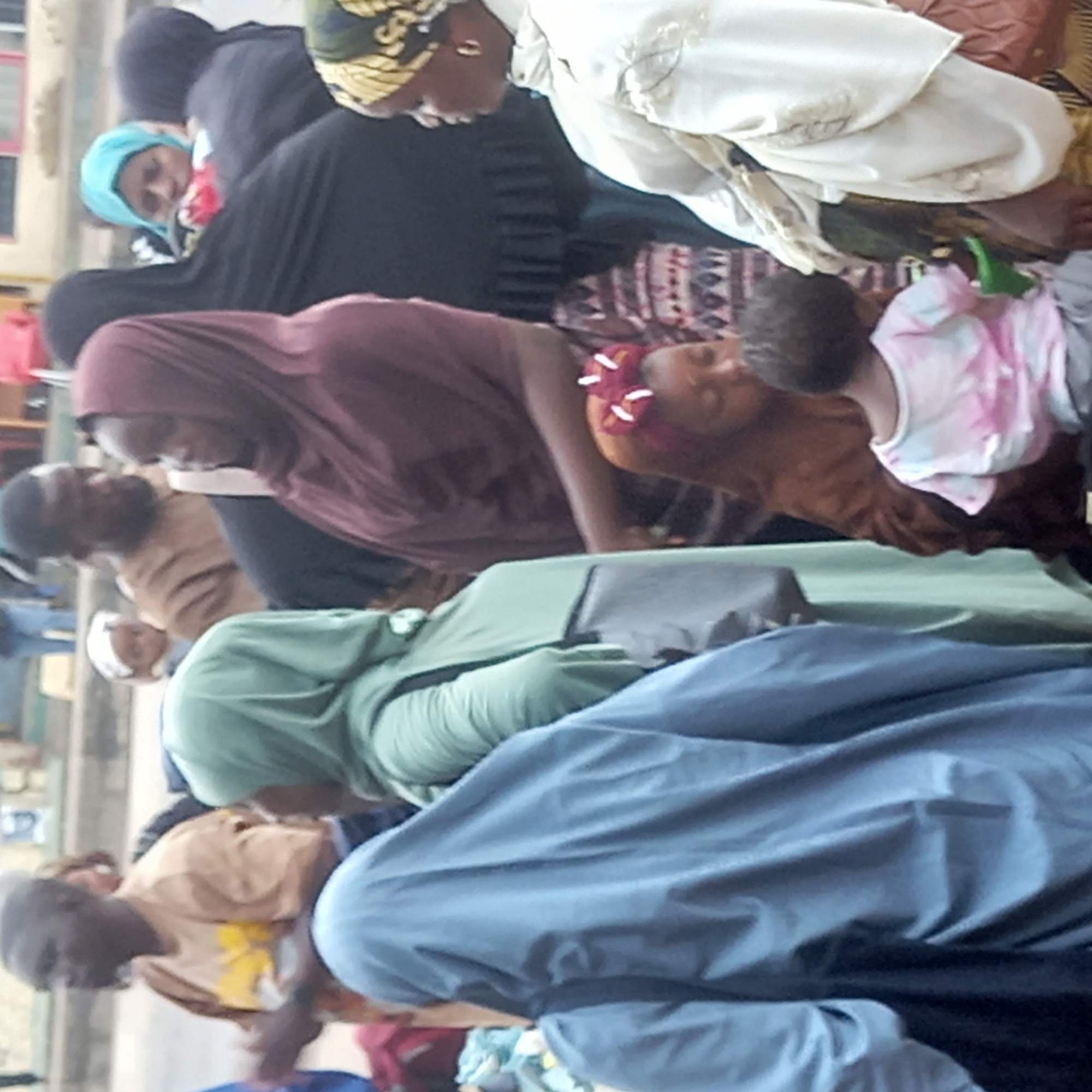 A street-level photo in Dadi Riba, Nigeria, shows a crowd of people. The individuals primarily appear to be women wearing hijabs or head coverings, clad in various fabrics and colors, including blue, green, brown, and black. Some are standing and conversing, while others seem to be seated. In the upper right, a child in a pink and white tie-dye shirt is visible. The environment is a mix of public space and buildings, with a wall visible in the background. The lighting suggests it is daytime.