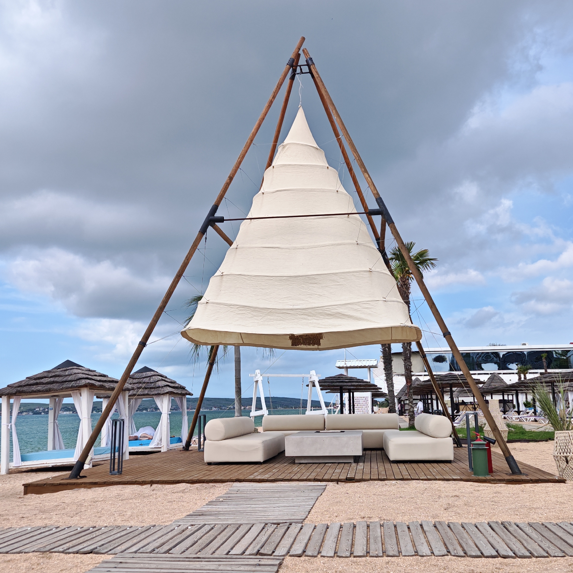 The image depicts a beachside lounge area in Мысовское сельское поселение, Ukraine. The focal point is a unique, teepee-style shade structure constructed from wooden poles supporting a layered, conical white canvas. Underneath this structure is a white modular sofa set with a gray concrete coffee table in the center. The entire setup is on a raised wooden platform. A wooden boardwalk extends towards the lounge area from the foreground. In the background, two white cabanas with thatched roofs and sheer white curtains stand on a similar wooden platform, with a person reclining on one of the daybeds inside. Further back, there is a white building and several thatched umbrellas, suggesting a resort or beachfront property. Palm trees and other greenery are also visible. The sky is overcast with a mix of gray and blue clouds. The ground is sand.