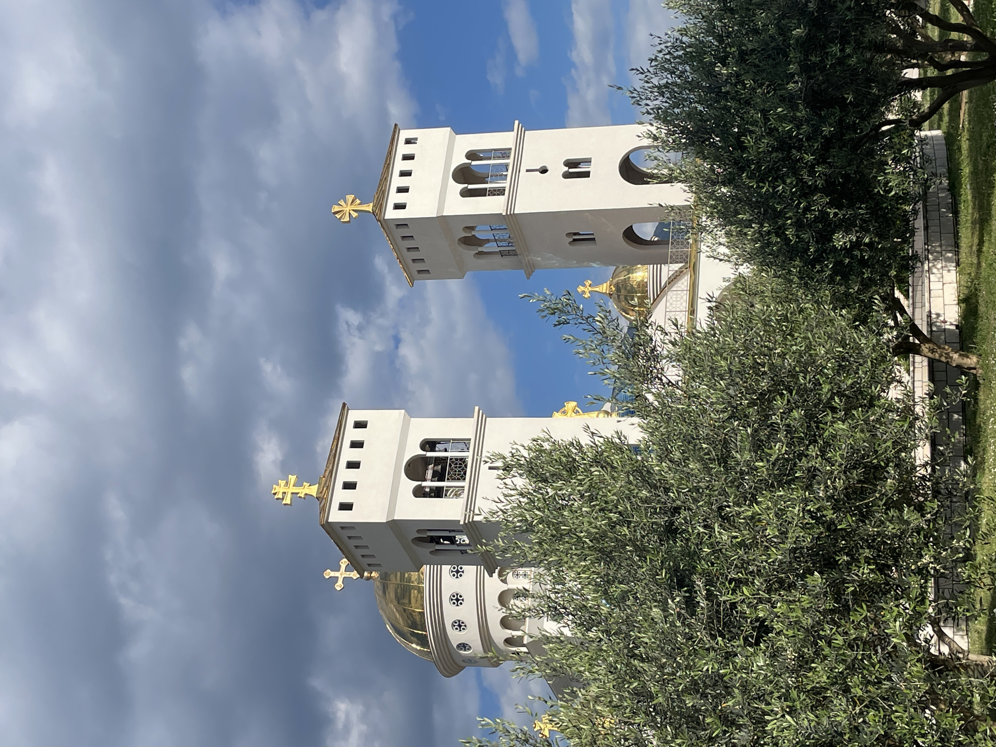 The image features a white Orthodox church building with gold crosses and a gold dome, partially obscured by trees. The church appears to have two towers with arched windows. The sky is cloudy with patches of blue. The location is likely Bar, Montenegro, given the architectural style of the building and the geographic context of the city. There are no people visible.