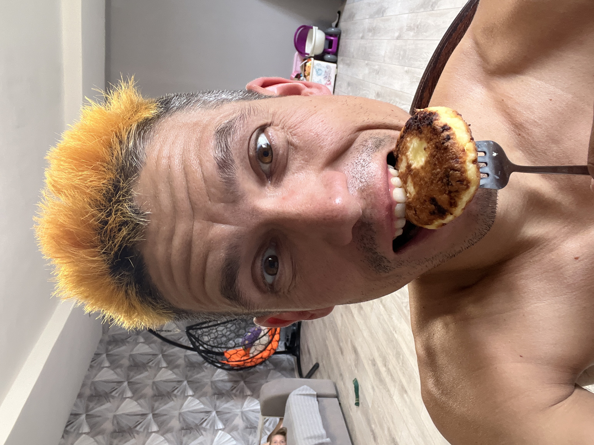 A close-up selfie of a shirtless man with spiked, bright orange-dyed hair is taken from the chest up. He is about to take a bite of a golden-brown, circular food item, possibly a pancake or fritter, which is held to his mouth on a fork. The man's mouth is open, exposing his teeth. His eyes are wide and looking directly at the camera. The background is blurred but appears to be an indoor domestic setting. To the left, a child is visible lying on a couch. To the right, toys, including a small purple and white stroller, are present. The flooring is light-colored wood. The image was taken in Chaikovsky, Russia.