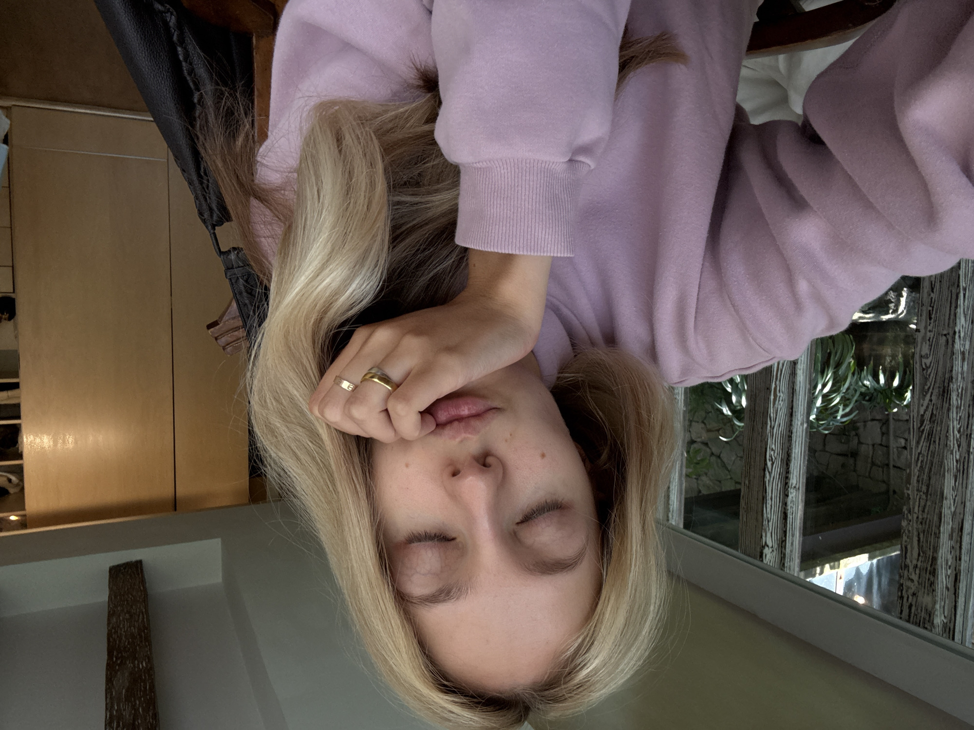 A young Caucasian woman with blonde hair and closed eyes is resting her chin on her hand, her lips pursed. She is wearing a pale pink sweatshirt and a gold ring on her right ring finger. The background shows a room with large windows, plants, wooden beams, and wooden cabinets. The overall impression is of a casual, indoor setting.