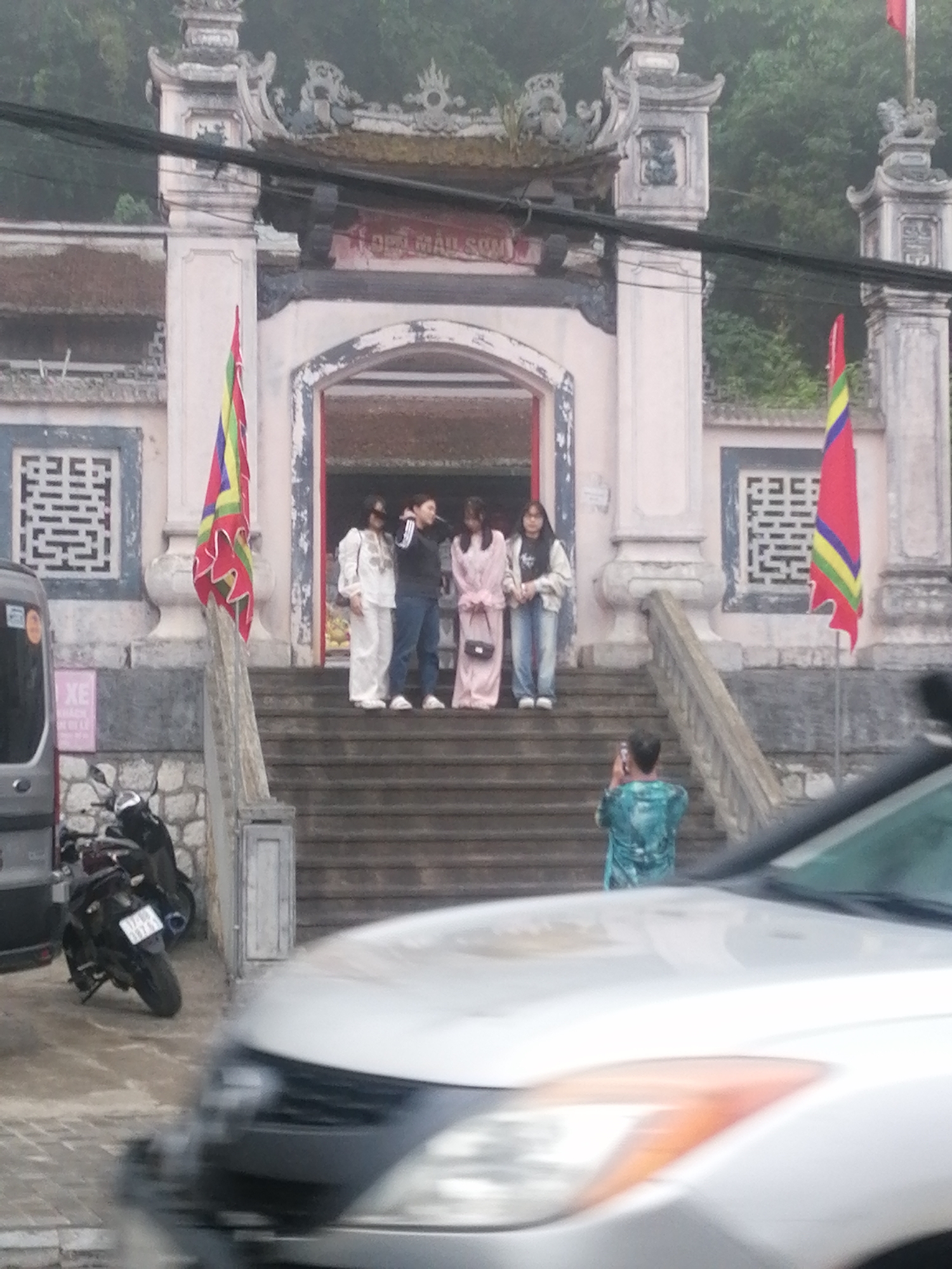 The image shows a group of four people posing for a photo on the steps of a temple entrance. The temple is of traditional Vietnamese design with ornate details and two Vietnamese flags flanking the entrance. A person is taking a photo of the group. In the foreground, a silver car and a motorcycle are partially visible. The location is Sa Pa Ward, Vietnam.