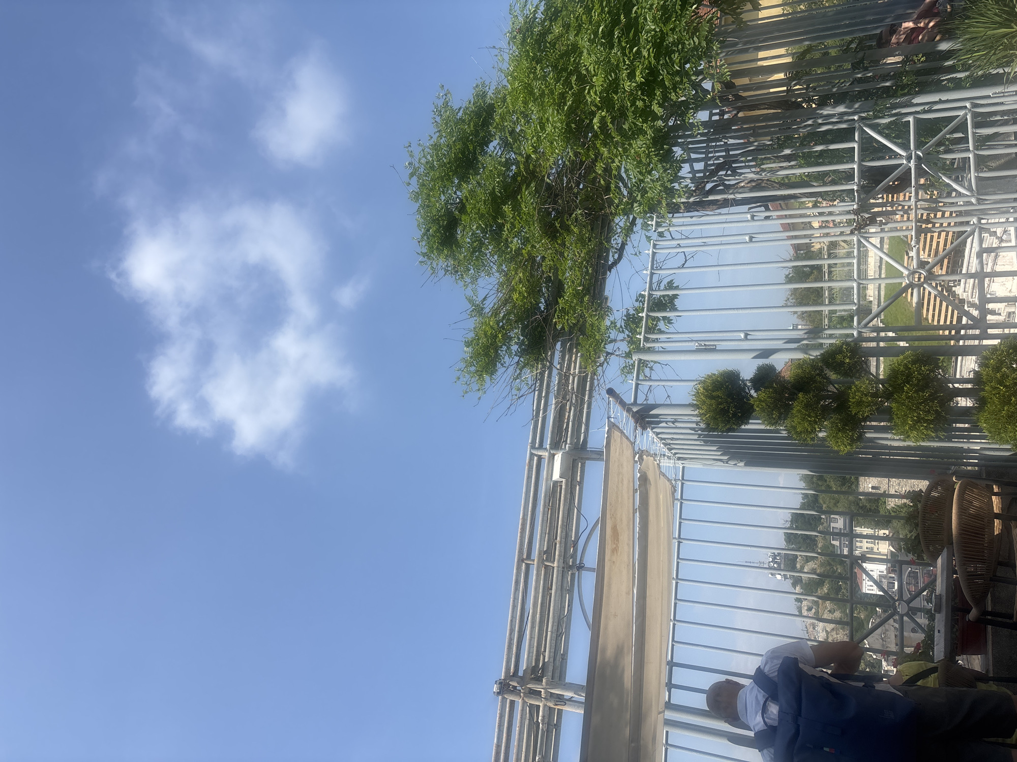 The image captures an outdoor scene, likely from an elevated terrace or balcony in Plovdiv, Bulgaria, on a bright, sunny day. A clear blue sky with scattered white clouds dominates the upper portion of the frame.
A prominent feature is a silver-grey metal railing or pergola-like structure, which extends horizontally across the image, framing the view. Attached to this structure is a light-colored, rectangular canopy or awning component.
In the foreground, towards the bottom right, a person is seated with their back mostly turned to the camera. They are wearing a white t-shirt and a blue backpack. To their right, parts of wicker or woven chairs are visible, suggesting an outdoor seating area, possibly belonging to a cafe or restaurant. A hint of another person's arm or shoulder can be seen behind the railing to the left.
Beyond the metal structure and railing, an abundance of green foliage and trees is visible, with a large, leafy tree occupying the upper left. In the distant background, urban buildings and structures spread out, with a faint antenna or tower discernible on a faraway hill, offering a panoramic view of the city. The overall impression is one of leisure and enjoying the view from an elevated vantage point on a pleasant day. There is no visible text in the image.