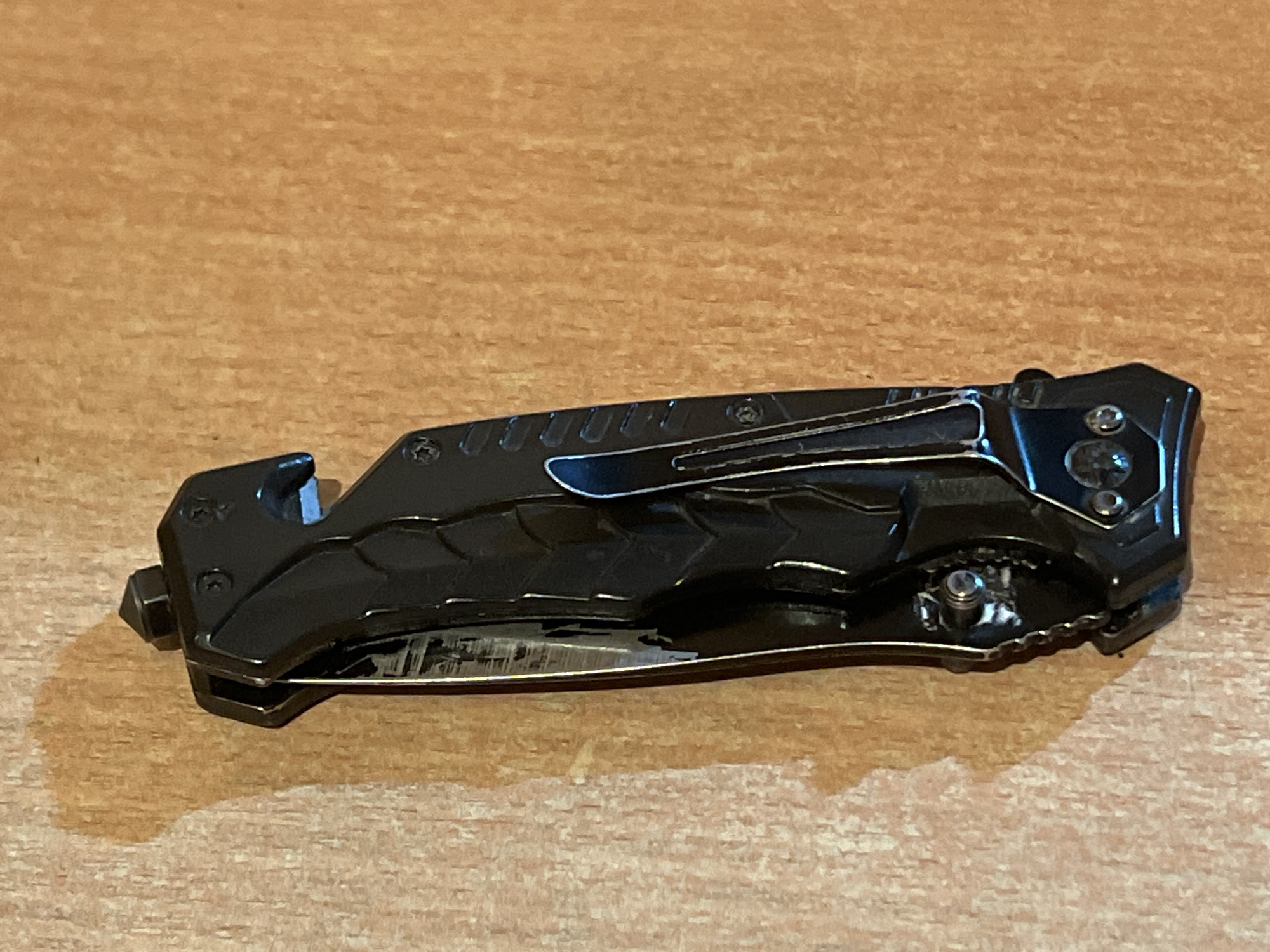 A black folding knife is lying on a wooden surface. The knife is closed and its blade is not visible. It has a clip attached to the side and appears to be made of metal. The wooden surface has a light brown, grainy texture. The image is taken from a top-down perspective. The scene is indoors, and there is a shadow cast by the knife on the surface. The location is specified as Iksha, Russia.