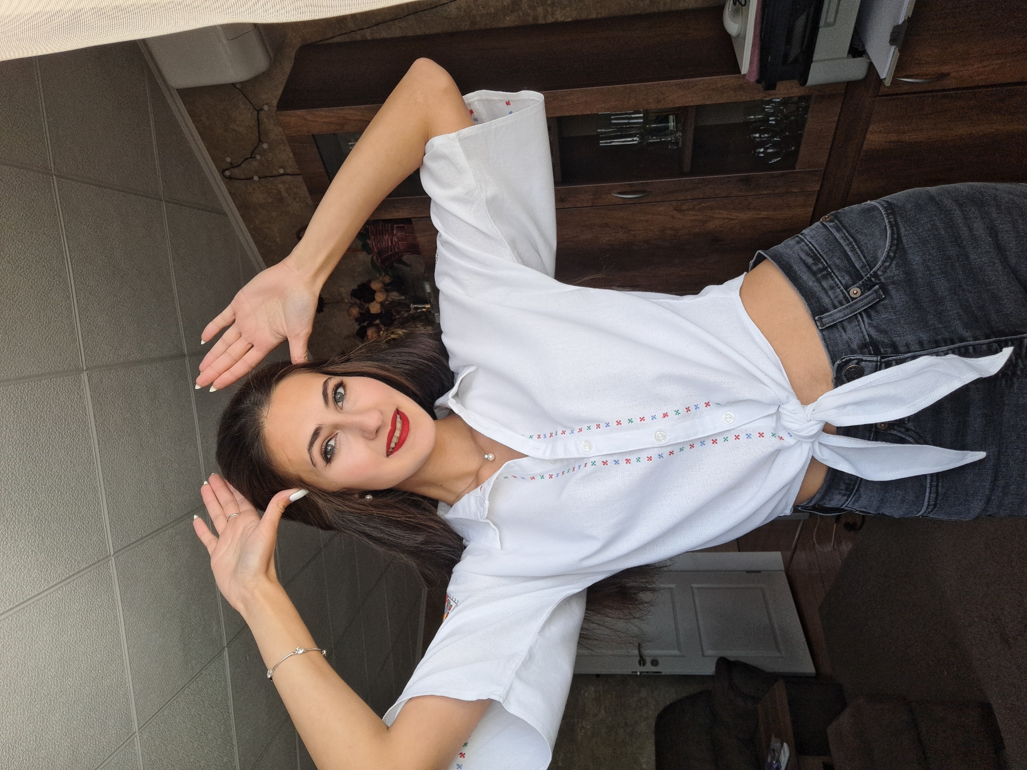 The image depicts a woman lying on her back, posing with her arms raised. She is wearing a white, button-down shirt with a tie at the waist and dark-wash jeans. The shirt is adorned with a decorative pattern along the button line. She has dark hair, fair skin, and red lipstick. Her right arm has a silver bracelet.
The background includes a tiled wall, a wooden cabinet, and a glimpse of an interior with what appears to be a door and seating area. The setting appears to be indoors.