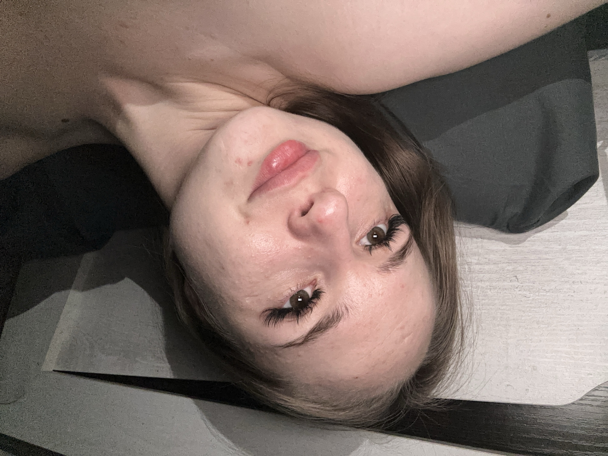 A young woman with brown hair and brown eyes is lying on her side, looking up and to the left. She is wearing false eyelashes and has a few blemishes on her face. Her lips are pink and slightly parted. The background consists of a white textured wall and a dark pillow. The lighting is dim, casting shadows on the wall and the woman's body.