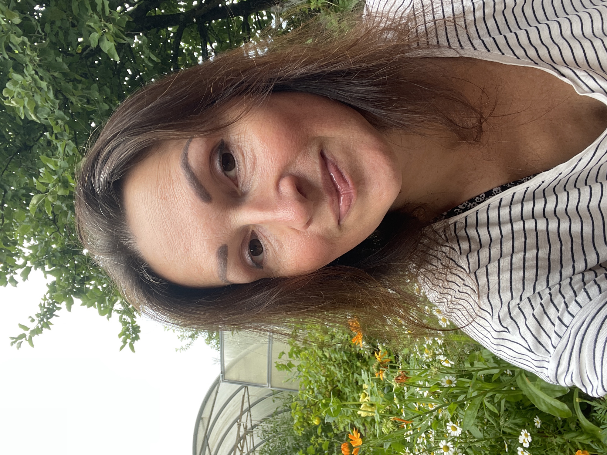 A woman with brown hair and brown eyes is taking a selfie outdoors. She is wearing a white t-shirt with black stripes. In the background, there are green trees, a greenhouse, and flowers. The scene appears to be in a garden.