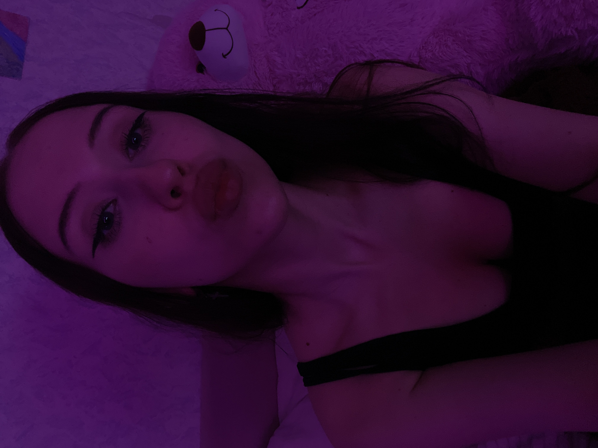 The image shows a young woman with long dark hair, wearing a black top, taking a selfie in a room illuminated by purple light. She is making a kissing face, and her makeup includes winged eyeliner. To her right, a large, fluffy pink teddy bear is partially visible. The background appears to be a wall with a textured surface and a framed artwork. There is no indication of the location being Votkinsk, Russia, or any specific city.