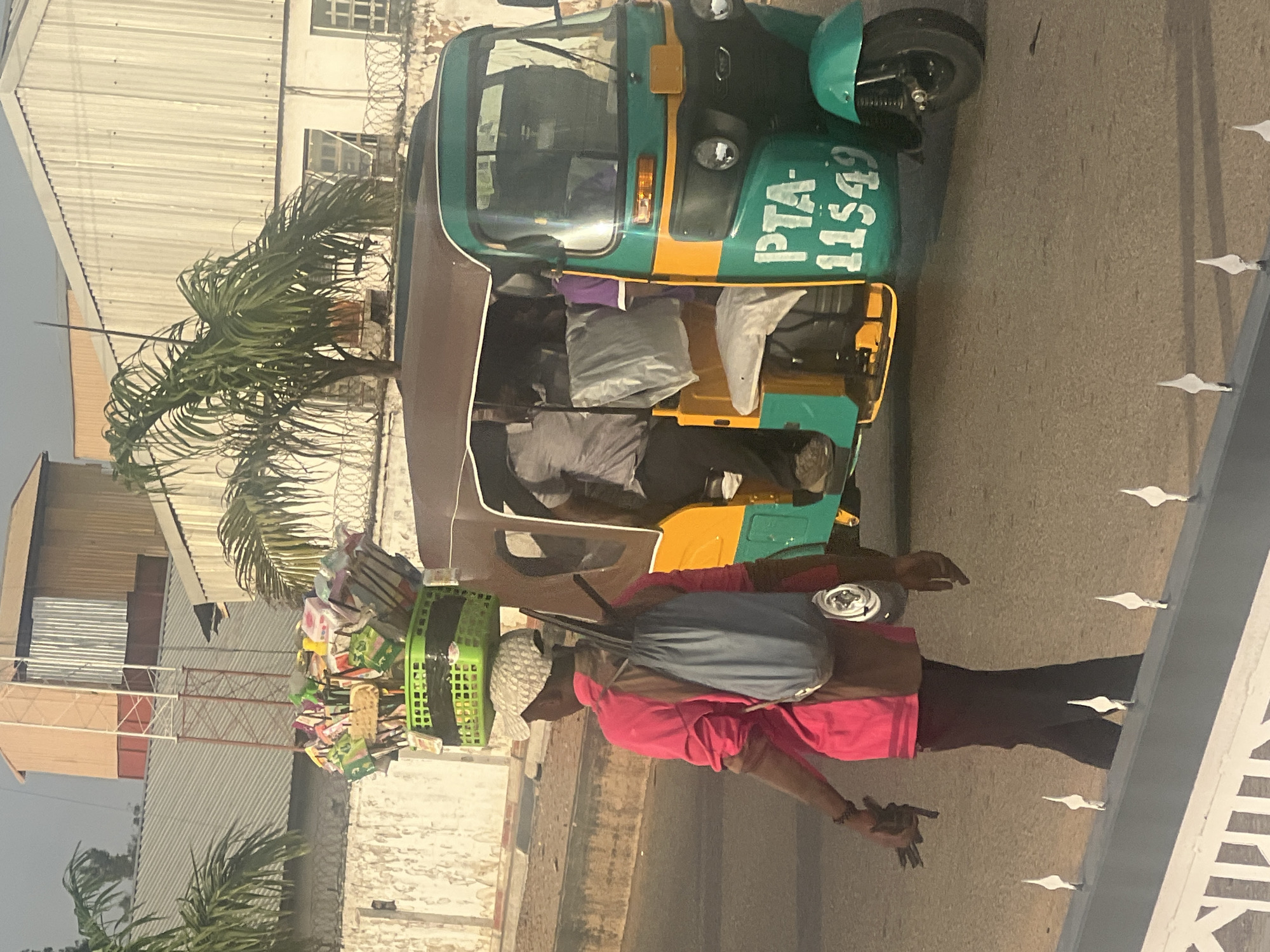 A medium shot depicts a person in a pink shirt and dark pants walking to the right of a green and yellow motorized rickshaw, with the license plate PTA-11549 visible on the side. The person carries a blue bag on their back and a collection of baskets and miscellaneous items balanced on their head. The rickshaw is parked on a paved surface, and passengers are visible inside. A palm tree is to the left of the rickshaw, with buildings and fences in the background. The setting is likely Barakin Lerawi, Nigeria.