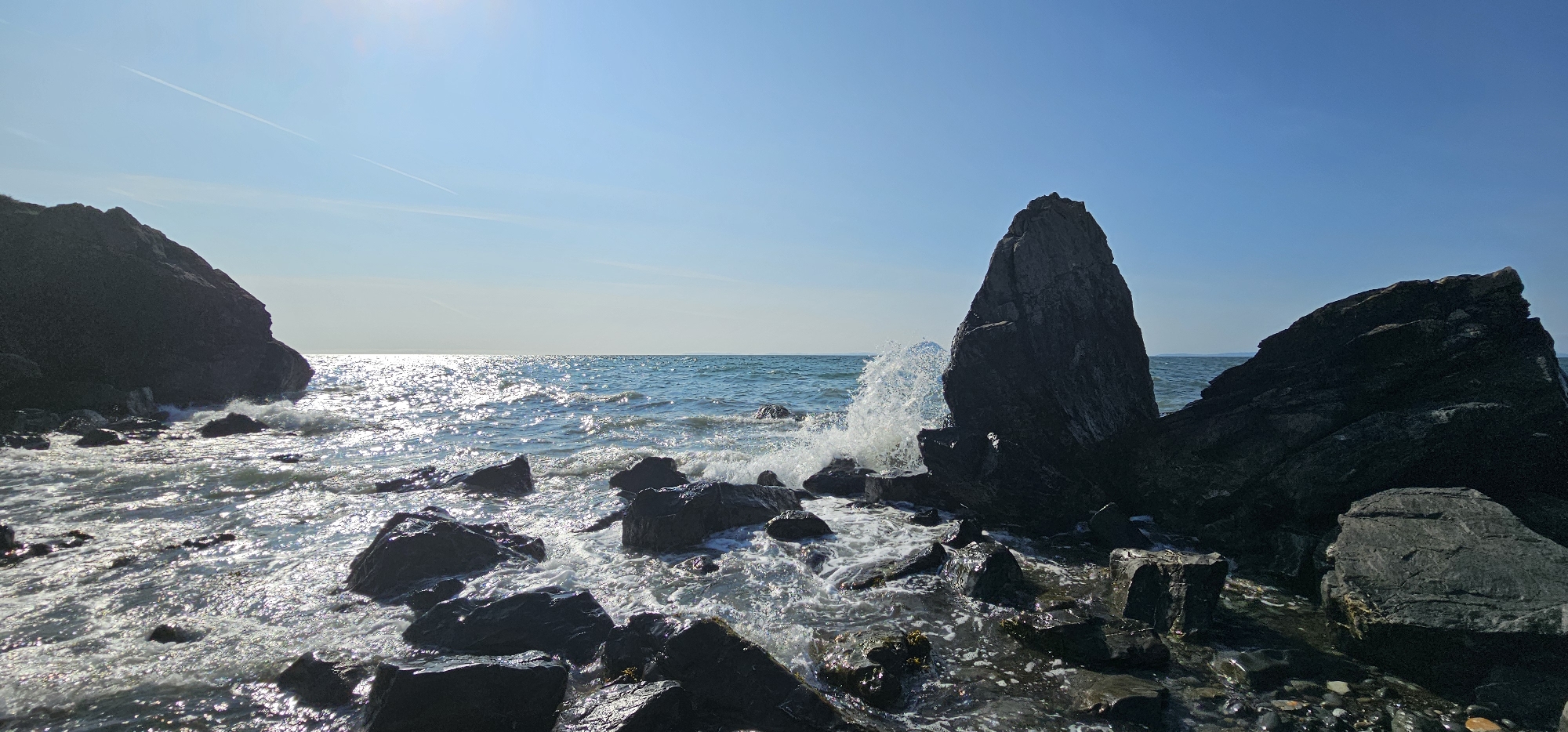 The image shows a rocky coastline on a clear, sunny day. The foreground is dominated by dark, wet rocks of various sizes, with waves crashing against them and creating white foam. In the mid-ground, the sea is a vibrant blue, sparkling with sunlight. A large, pointed rock formation stands prominently in the right mid-ground, with a wave breaking against its base. In the background, the sea meets the horizon under a clear, pale blue sky. Two faint white contrails are visible in the sky. The scene depicts a natural coastal landscape.