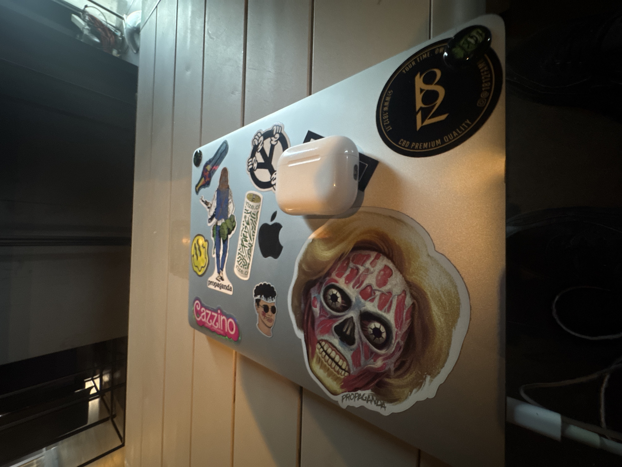 This is a close-up, slightly overhead shot of a silver laptop placed on a light-colored, slatted wooden surface. The laptop is adorned with several stickers. A prominent sticker on the left features a stylized, anatomically detailed skull with blonde hair, labeled "PROPAGANDA." To its right, there's a smaller sticker of a face with dark curly hair and sunglasses. Other stickers include a yellow smiley face, a bird, a peace sign with hands, and a sticker with text. Resting on the laptop is a white wireless earbud case. On the bottom right of the laptop is a round black sticker with gold lettering that reads "CBD PREMIUM QUALITY" and "YOUR TIME OWN." The background is dark and out of focus, with what appears to be furniture and window blinds visible. The scene suggests a casual setting, possibly a cafe or home, and no people are visible. The image is well-lit, highlighting the stickers and the laptop.