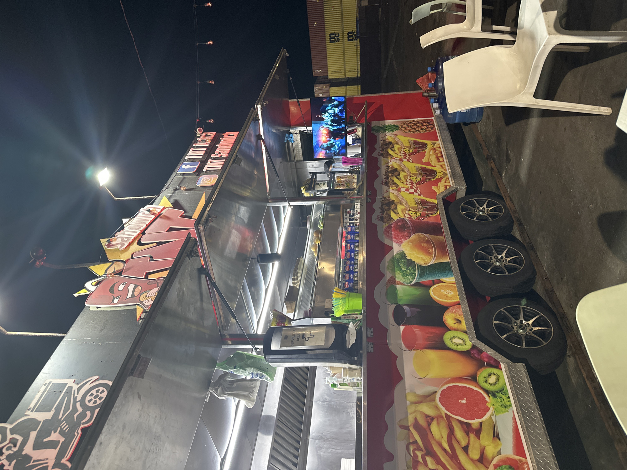The image depicts a food truck, possibly in the city of Arica, Chile, given the writing on the signage. The truck features a large, illuminated sign with the name of the establishment. The side of the truck is decorated with images of various foods, including fruits, fries, and possibly other snacks. A television is mounted on the side of the truck displaying a video. Several white plastic chairs are visible near the truck. The surrounding environment appears to be an outdoor area, possibly a parking lot or event space, with storage containers in the background. The photograph was taken at night.