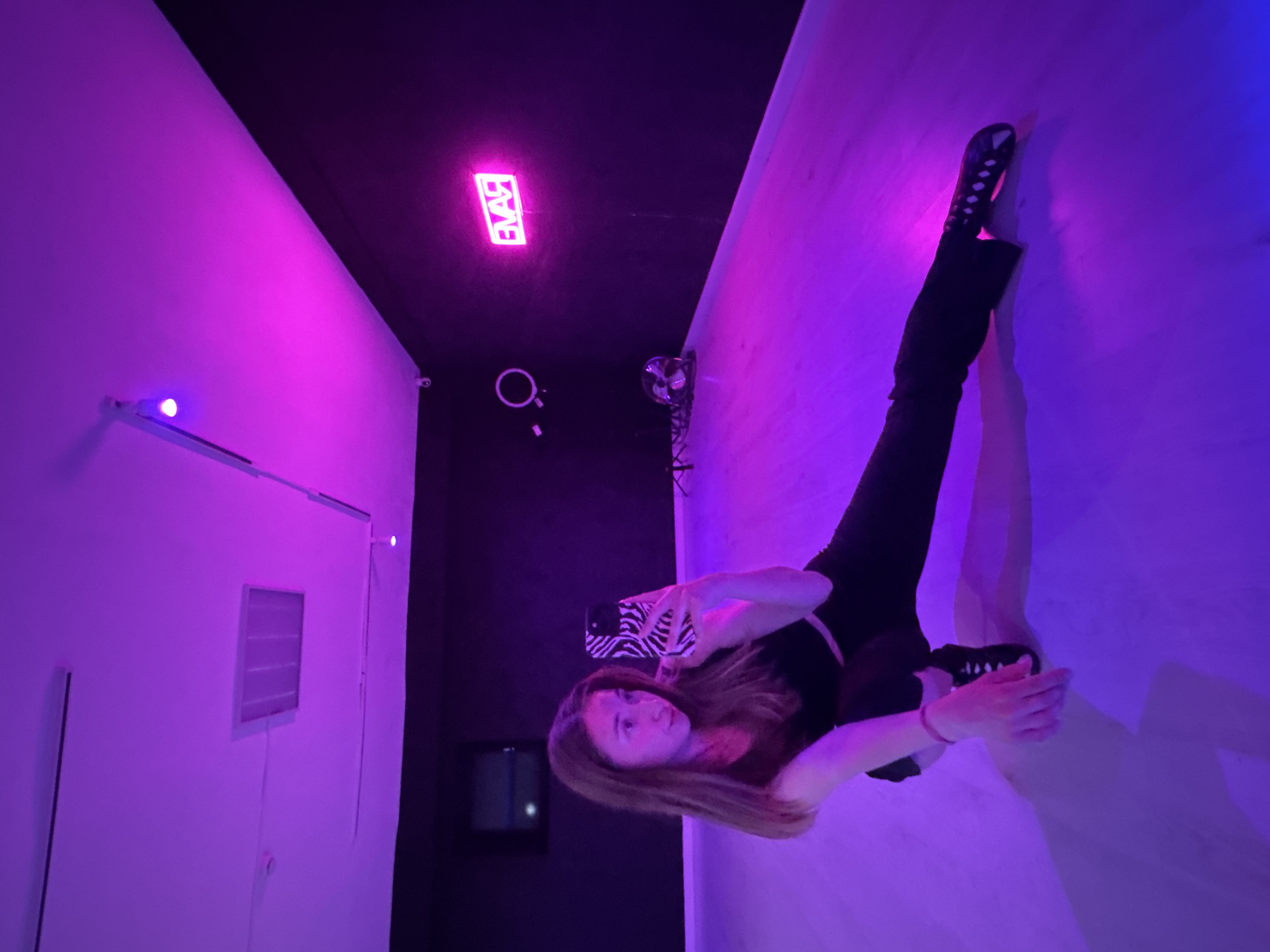 A young woman is taking a selfie in a room with purple and blue neon lighting. The room has dark walls and ceiling, with a "EVAR" neon sign on the ceiling. The woman is wearing dark clothing and sneakers. The exact location in the city of Chaikovsky, Russia, is not discernible from the image.