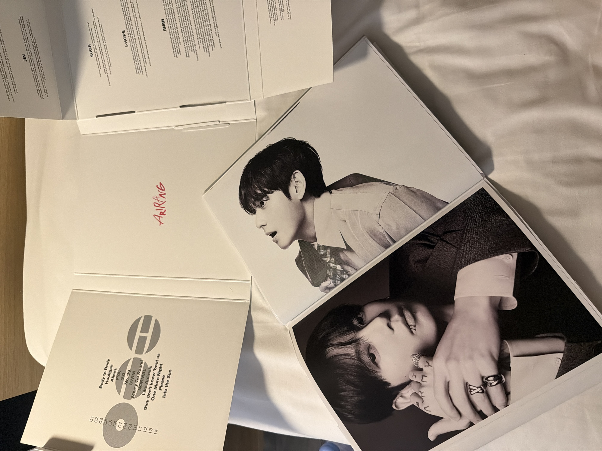 An open white box lies on a white bedsheet. The box has a title in red cursive font that reads "ARIRANG". Inside the box, on the left, are song titles and track numbers, with the number "07" highlighted in a gray circle. To the right, the box is divided into three sections. The first section contains text in a small font with "JIN" written above it. The second section has "SUGA" written above text in a small font. The third section has "J-HOPE" written above text in a small font. There is also a loose booklet open in front of the box. The booklet has a black and white photograph of a man's face on the right page. The left page of the booklet shows a close-up of a man's hands with rings on his fingers, and tattoos on his arm. The overall scene is of merchandise, possibly a music album or photo book, laid out on a bed. There is no indication of the location being in Ivry-sur-Seine, France.