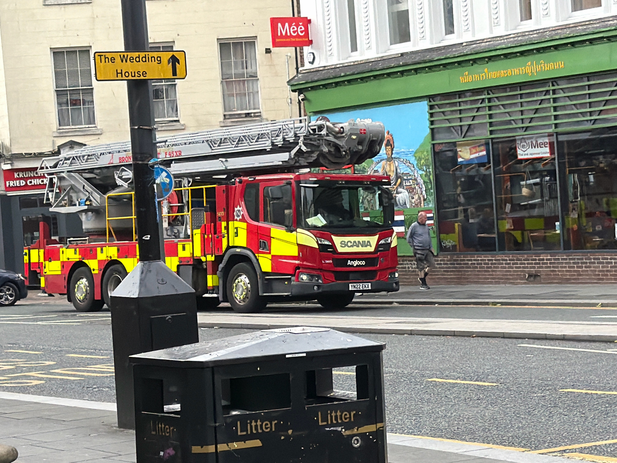A red and yellow fire engine is parked on a city street in front of a building. The fire engine has "SCANIA" and "ANGLOCO" written on the front. A sign on a pole above the fire engine reads "The Wedding House" with an arrow pointing up. To the right of the fire engine, a man in a grey shirt and shorts walks away from the camera. The building behind them has a green facade with windows and a mural. A red sign on the building reads "Mee". Two black trash bins are in the foreground. The street is paved and marked with yellow lines.