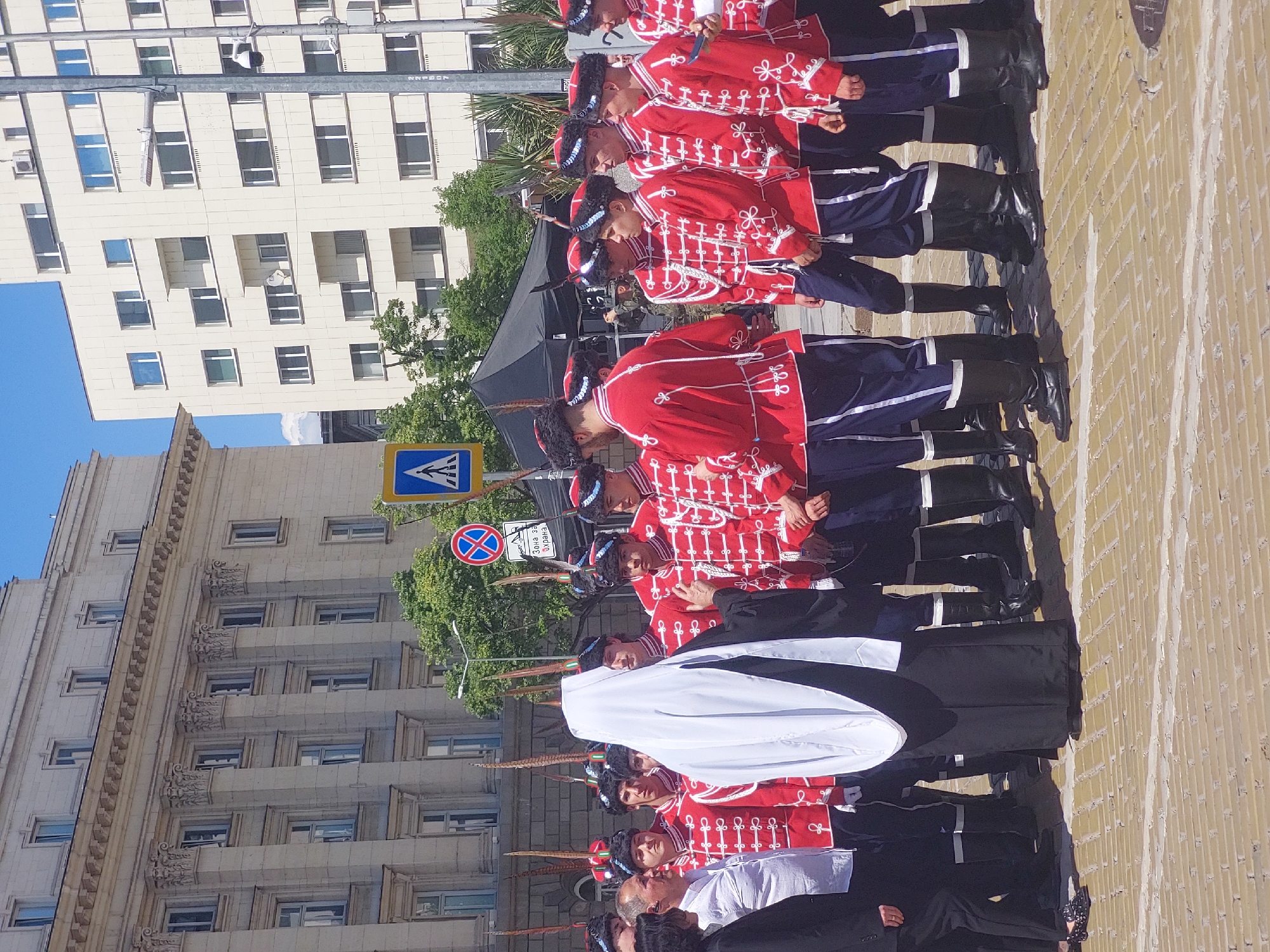 Here's a detailed description of the image:
**Scene Overview:**
The image shows a line of soldiers in distinctive red and navy blue uniforms, standing at attention on a brick-paved area. Behind them are several large buildings, one of which appears to be a modern office building with many windows, and another of older architecture. The sky is clear and blue.
**People:**
The soldiers are arranged in a neat row. They wear red jackets with white detailing, navy blue trousers, and tall black boots. They have hats adorned with feathers. Several individuals in black formal wear are interspersed among the soldiers. A white drape or flag is held by several soldiers toward the front of the line.
**Objects:**
The buildings in the background provide the urban setting. Street signs and trees are also visible. The brick pavement forms the foreground.
**Actions & Interactions:**
The soldiers are standing in a formal military formation, likely during a ceremony or performance. The people in black formal wear could be officials or guests. The white drape suggests a formal function.
**Environment:**
The scene is located in an urban area, with modern and classical architecture indicating a historical or administrative center. The bright daylight suggests a sunny day.