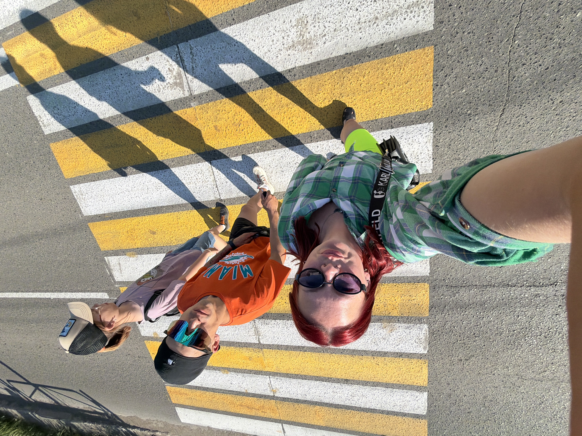 A high-angle group photograph shows three individuals on a pedestrian crossing with alternating white and yellow stripes. The location is likely Chaikovsky, Russia, based on the user's prompt.
The first person is wearing a green and white plaid shirt, bright green shorts, black shoes, and black sunglasses. They are holding the camera. The second person is wearing an orange "MIAMI" t-shirt, black shorts, sunglasses, and a black baseball cap. The third person wears a tan and black trucker hat, a light purple t-shirt, and light-colored jeans.
The shadows of the individuals are cast onto the pedestrian crossing. The crossing is on a grey asphalt road.