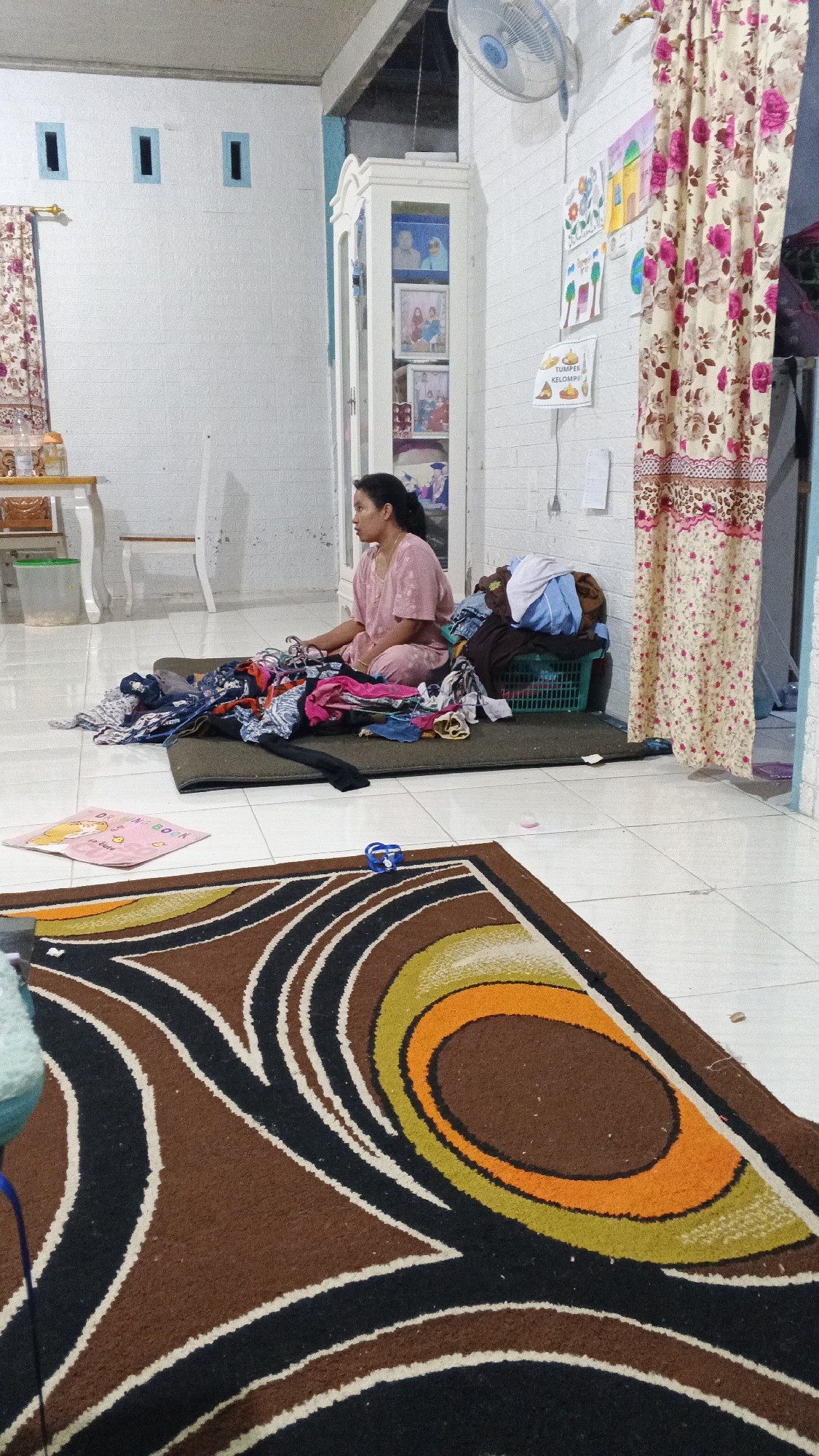 In Kampung Rempak, Indonesia, a woman is seated on a dark mat on a white tiled floor, surrounded by a pile of unfolded clothes. She is wearing a pink shirt and pants. To her right, a green laundry basket contains more folded clothes. Behind her, a white cabinet displays framed pictures. The room is lit by artificial light, and a ceiling fan is visible. The walls are white, with some decorative elements and a window with blue shutters on the left. A patterned rug is in the foreground, partially obscuring a drawing book. Curtains with floral patterns hang on the right side of the image.