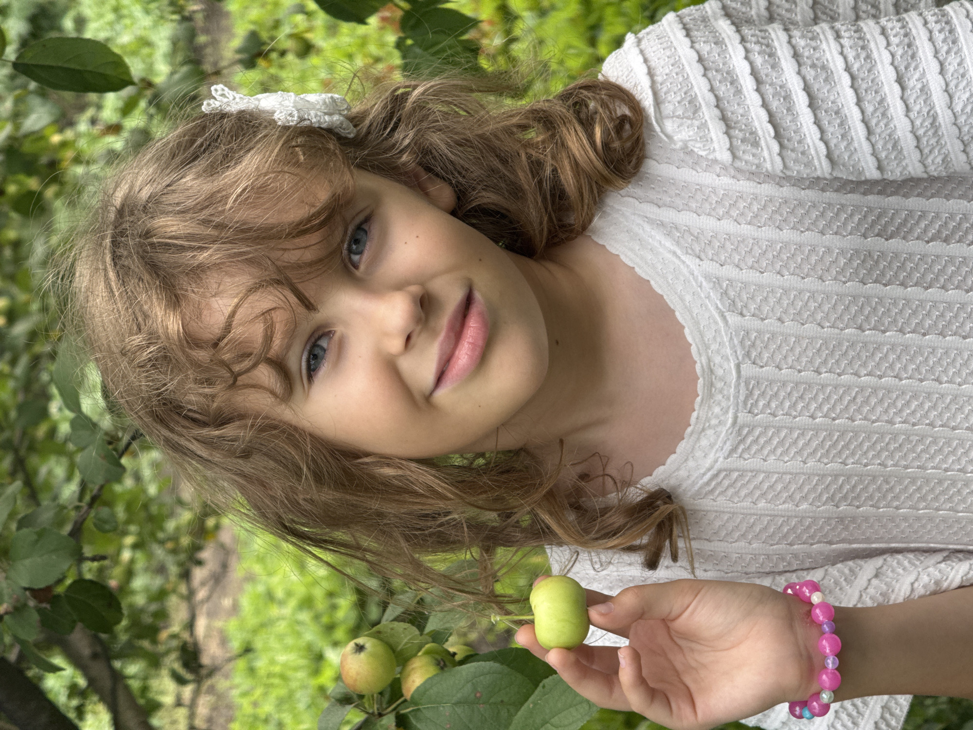 A young girl with curly blonde hair and blue eyes, wearing a white textured dress and a pink beaded bracelet on her left wrist, stands outdoors amidst lush green foliage. She is holding a small, green apple in her left hand. The background consists of blurred green leaves and trees, suggesting a natural outdoor setting, possibly a garden or orchard. The lighting indicates it is daytime.