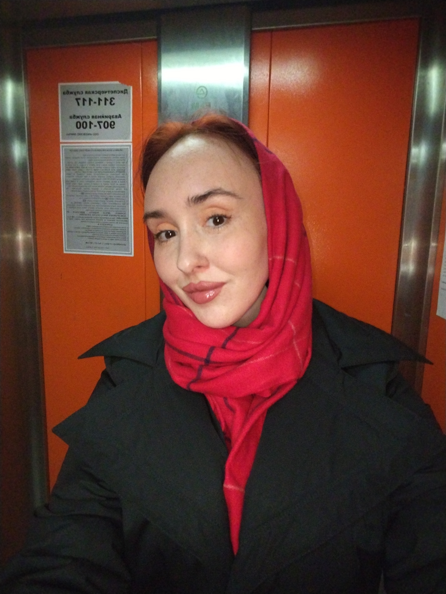 A fair-skinned woman with auburn hair and wearing a red headscarf and dark coat stands in an elevator with orange interior panels. A sign in Cyrillic is visible to the left of her head. The location is Izhevsk, Russia.