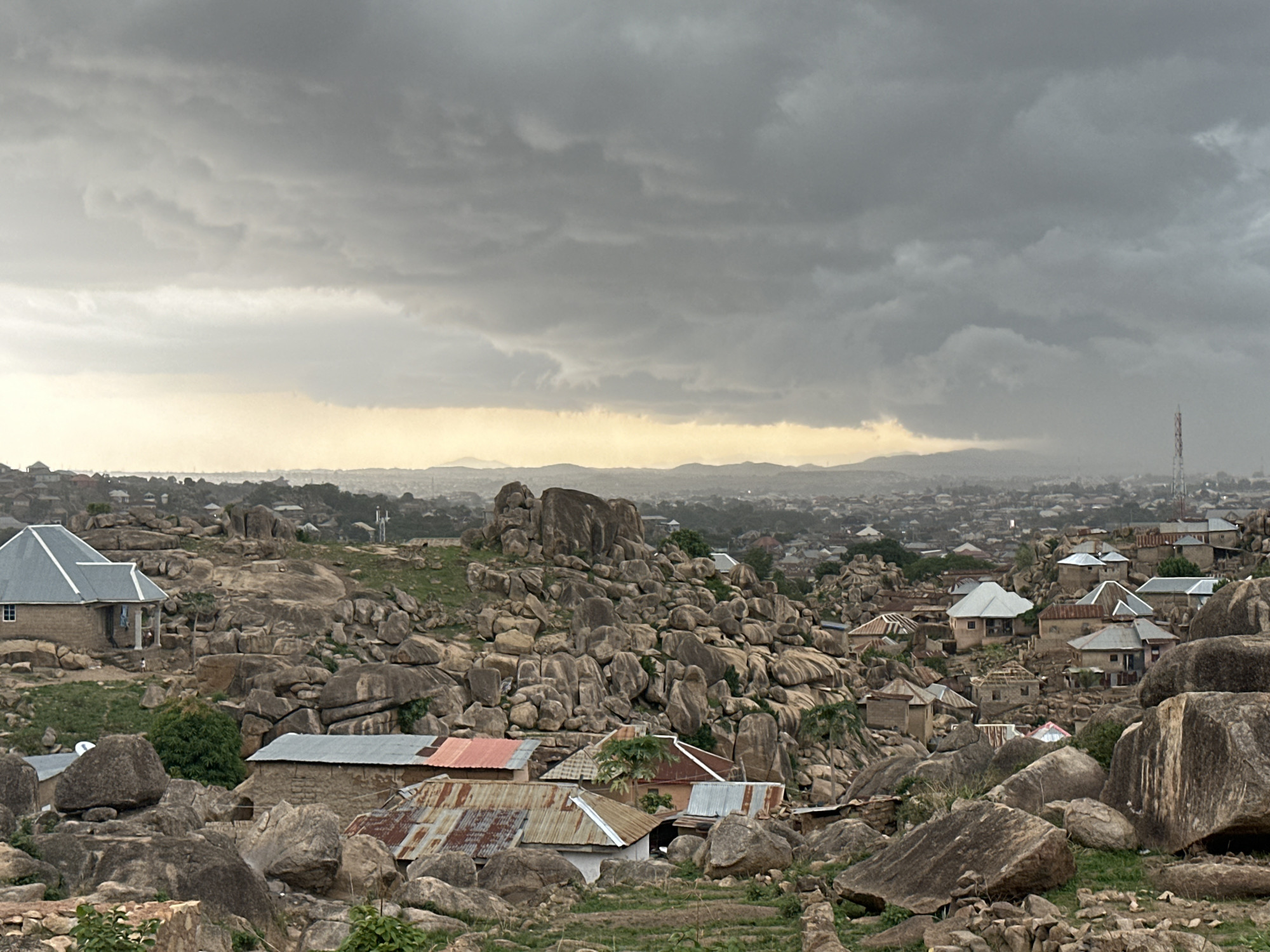 The image shows a residential area in Jos, Nigeria, under a cloudy, overcast sky. The landscape is characterized by large, rounded granite boulders interspersed among the buildings. The houses are a mix of single-story structures with corrugated metal and tile roofs. In the background, the city stretches out toward the horizon, disappearing into the hazy atmosphere. A telecommunications tower is visible on the right side of the image. There are no people visible in the image.