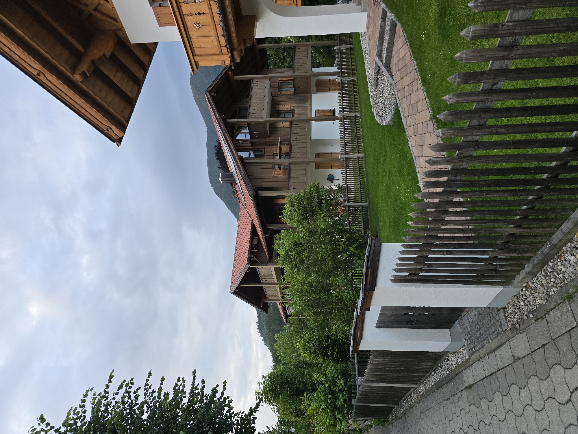This image depicts a scene in Garmisch-Partenkirchen, Germany. The primary focus is on a traditional Bavarian-style house with a red tiled roof and wooden balconies. The house is surrounded by lush green lawns, dense trees, and a rustic wooden fence. A paved walkway runs along the side of the property, adjacent to the fence. In the background, rolling hills and mountains are visible under a cloudy sky. The architectural style suggests a residential property in a scenic, possibly mountainous, region. There are no people or visible actions taking place; the scene is static and captures the exterior of the property and its natural surroundings.