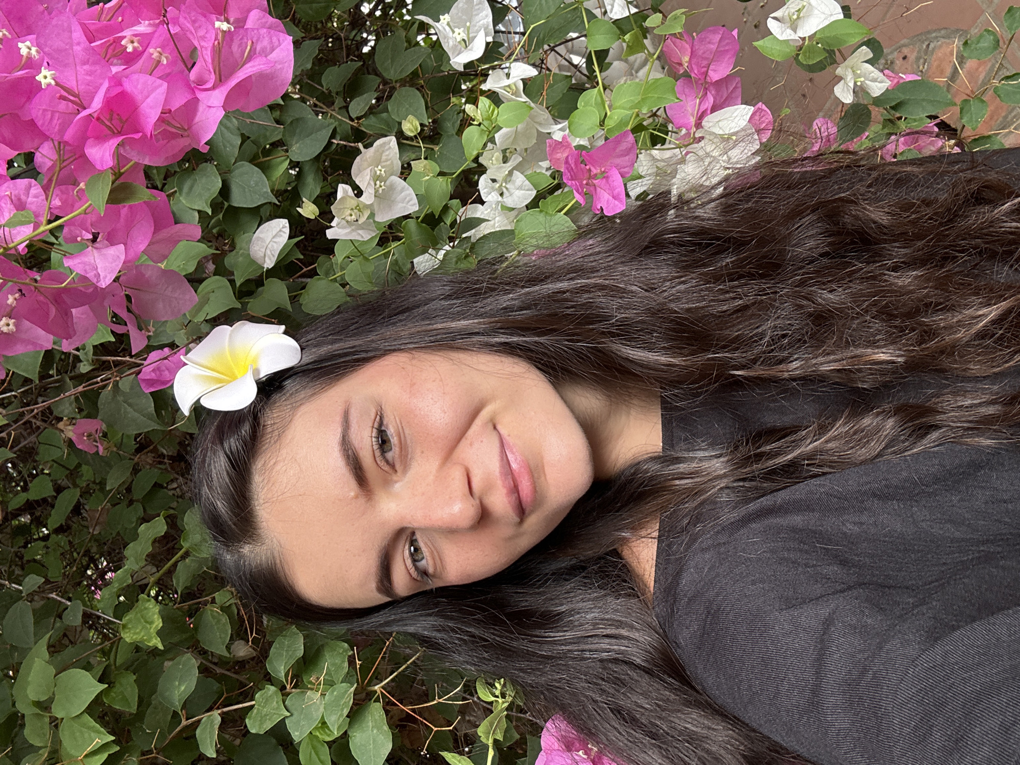 A woman poses for a portrait outdoors, likely in a garden or natural setting. She has long, wavy, dark brown hair and a white and yellow frangipani flower tucked behind her ear. She is wearing a black shirt. The background is filled with vibrant bougainvillea flowers in pink and white, along with green foliage. The photo is taken in Mũi Né, Vietnam, as indicated by the tropical flora.
