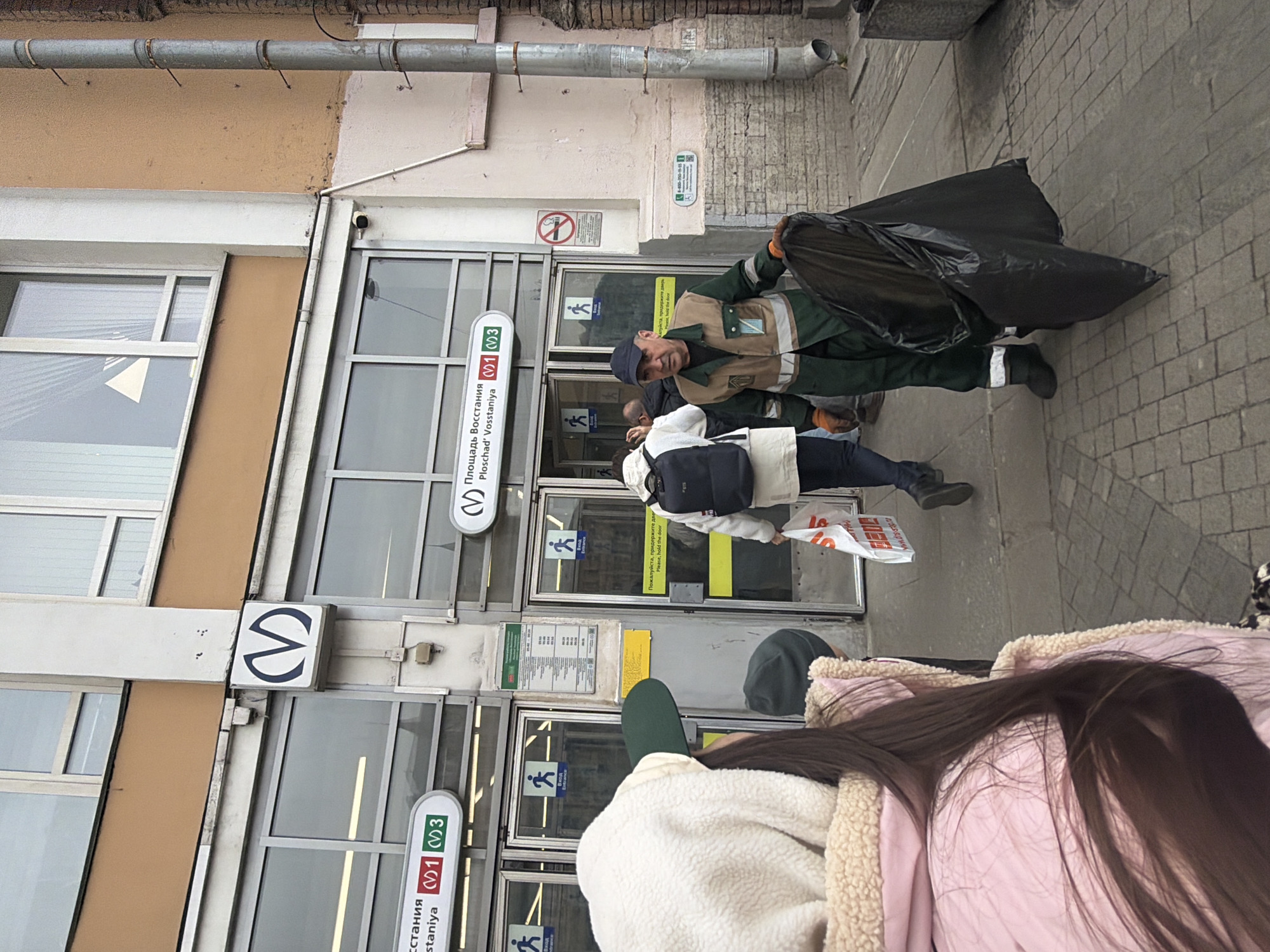 An outdoor scene depicts the entrance to the Ploshchad Vosstaniya (Площадь Восстания) metro station in Saint Petersburg, Russia. The station entrance features a gray facade with multiple glass doors, marked with "ВХОД" (Entrance) signs and walking person icons. Above the doors, a horizontal sign displays "Площадь Восстания / Ploschad' Vosstaniya" and "V1 M3," indicating metro lines. A prominent blue 'M' logo for the metro system is visible on a white square sign.
A male sanitation worker or janitor, wearing a dark blue baseball cap, a green and tan uniform vest with reflective stripes, and orange gloves, stands in the foreground-midground holding a large black plastic trash bag in his hands, facing slightly left of the camera. To his left, another individual with their back to the camera, wearing a white puffer jacket and a black backpack, is entering the metro station while carrying a white plastic shopping bag with red text.
On the far right, a vertical gray downspout is affixed to an adjacent building wall, which is light beige and textured. A red circular "No Smoking" sign is mounted on the wall to the right of the metro entrance. The ground is paved with gray interlocking pavers. In the immediate foreground, a person with long brown hair, seen from behind, wears a white fleece hood, a dark green baseball cap, and a pink puffer jacket with a beige sherpa-like lining.