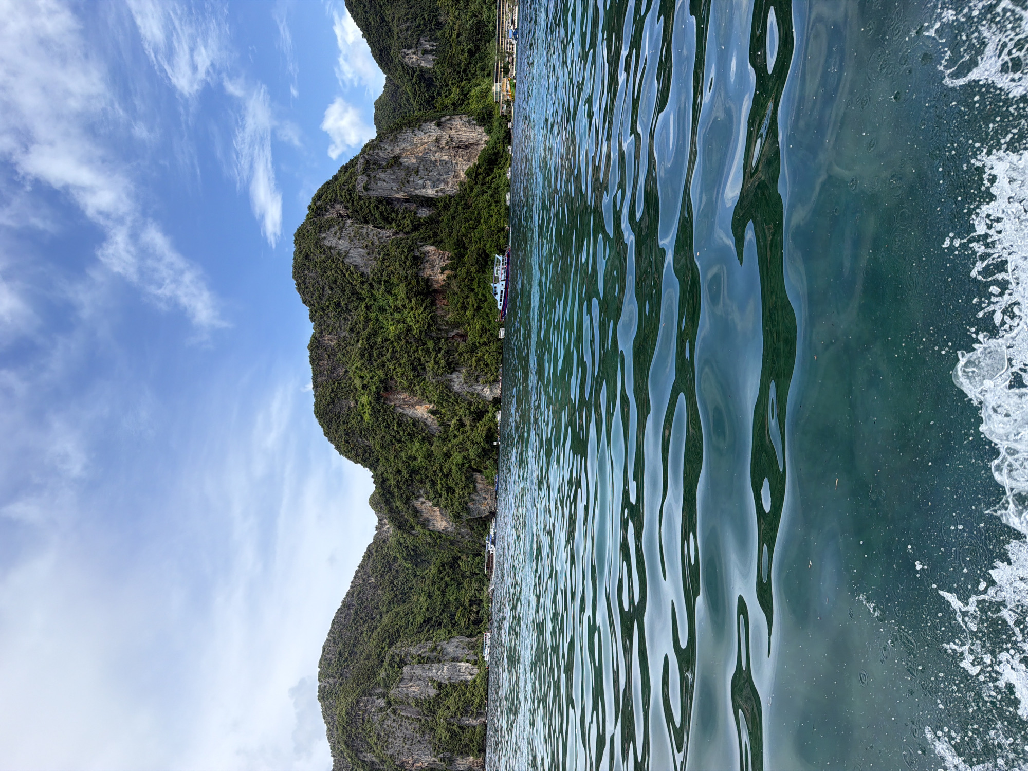The image depicts a view of Ao Nang, Thailand, from the water. In the foreground, the emerald green ocean is choppy with white foam generated by a boat's movement. The water surface reflects the sky and the surrounding limestone cliffs. In the middle ground, several long-tail boats and other vessels are anchored or moving in the water. The background features lush green, rocky cliffs that rise sharply from the sea. The sky above is a bright blue with scattered white clouds. A few buildings and structures are visible along the coastline at the base of the cliffs.