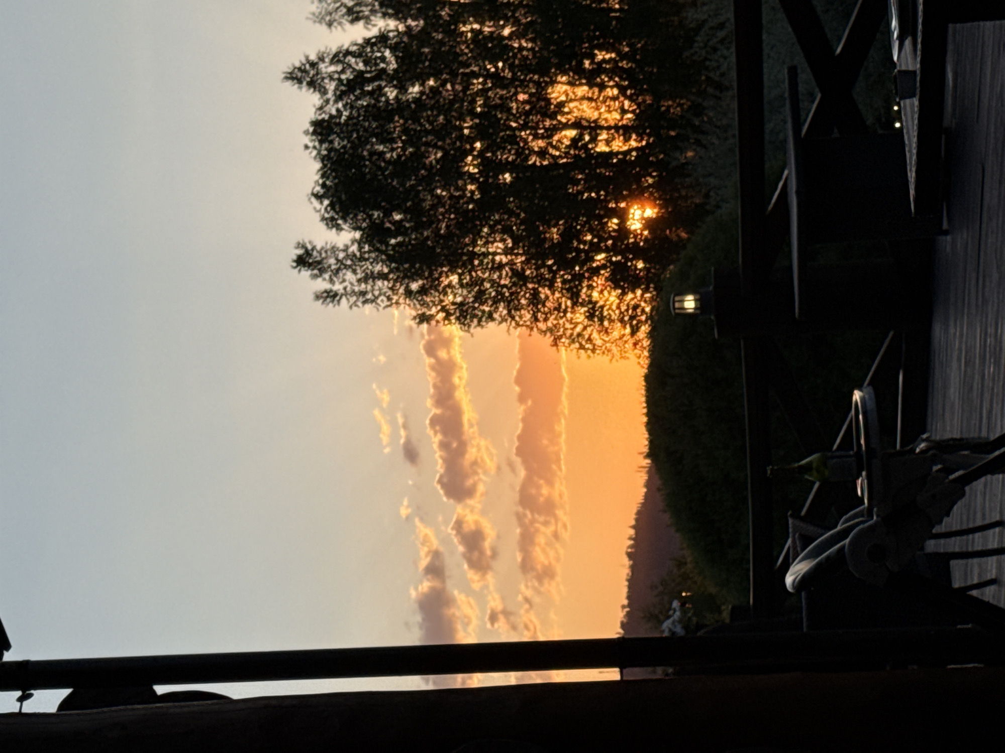 The image captures a serene sunset or sunrise over a natural landscape in Urlu-Aspak, Russia, observed from a wooden deck or balcony. The sky dominates the upper portion of the frame, displaying a gradient from a soft, pale blue on the left to a vibrant orange and yellow towards the right, indicative of the early morning or late evening. Several elongated, glowing cumulus clouds, illuminated from behind by the sun, stretch horizontally across the middle-left of the sky.
A large, dark tree with dense foliage is silhouetted against the radiant sky on the right side of the image, with glimpses of the bright sun peeking through its leaves. Below the sky, a dark, undulating horizon line suggests distant hills or a dense forest.
In the foreground, a dark wooden deck or balcony structure frames the bottom and right edges of the composition. A thick, dark wooden railing runs horizontally across the bottom. On the right, wooden beams form part of a railing or support, exhibiting criss-cross patterns. A lantern-style light fixture, emitting a warm glow, is attached to this structure. Further into the deck area, a chair with a light-colored cushion and a bottle (possibly green) resting on a surface are visible in silhouette.
The scene conveys a sense of peacefulness and natural beauty, offering a quiet vantage point to appreciate the shifting light of day. No visible text is present in the image.