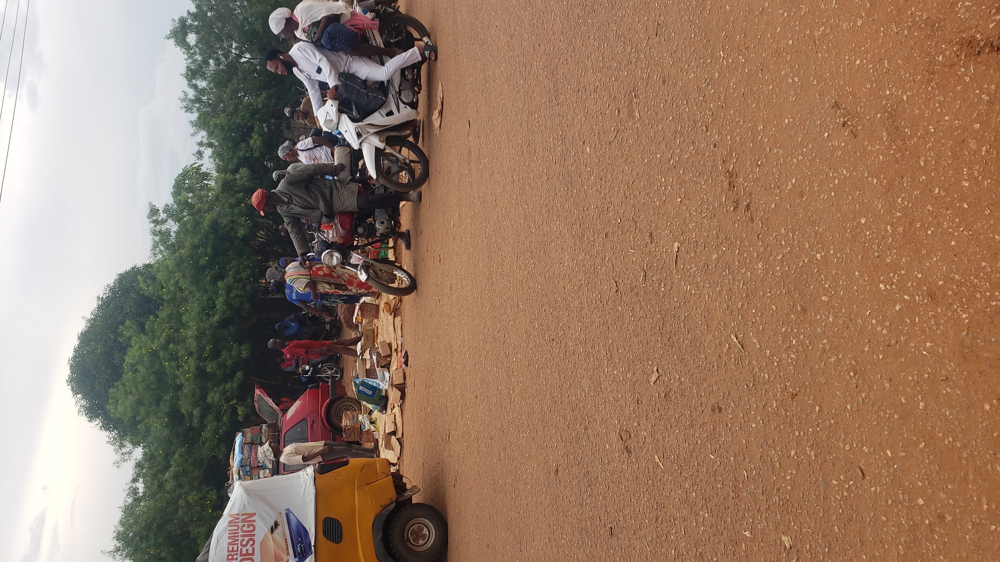 In Kakuri Gwari, Nigeria, an outdoor marketplace scene unfolds. A yellow three-wheeled auto rickshaw, branded with "PREMIUM DESIGN," is parked on the left. A red car with its trunk open is behind it, loaded with sacks. People are gathered around, some standing, others on motorcycles. One person on a red motorcycle appears to be talking to a person on a white motorcycle. Items are spread out on the ground, possibly for sale. The background features lush green trees under a cloudy sky. The ground is a reddish-brown, typical of Nigerian soil.