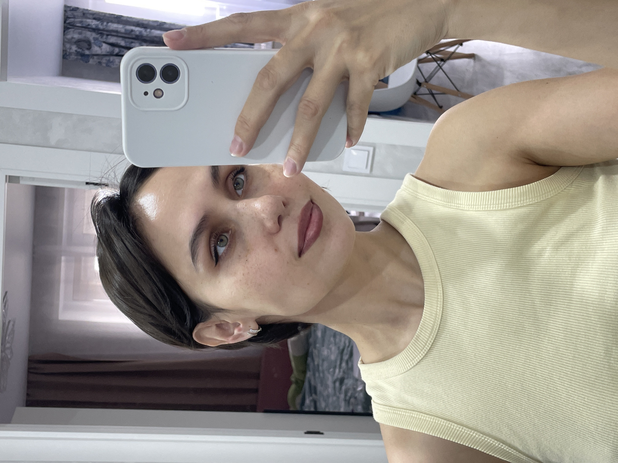 A young woman with short dark hair and light skin is taking a selfie in what appears to be an indoor location, potentially a home. She is holding a white smartphone with a dual-lens camera up to her face. She is wearing a light yellow ribbed tank top. The background is slightly blurred, but shows elements like a window with a curtain and a glimpse of a white chair. There is no information to conclusively determine if the location is the city of Izhevsk, Russia.