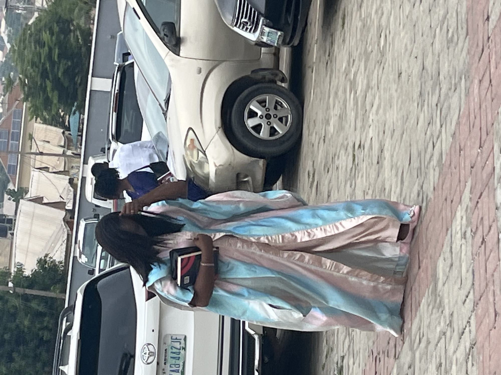 A full-body, eye-level shot captures a woman walking in what appears to be a parking lot in Abuja, Nigeria. She is wearing a flowing, multi-colored robe with pastel stripes of pink, blue, and white, and pink ballet flats. In her hands, she holds a closed book, a tablet, and what looks like a wallet or clutch. Her hair is dark and falls to her shoulders, and she is looking down.
Behind her, a white Toyota SUV is parked, with a license plate reading "YAB-442JF". Further in the background, other vehicles are visible, including a light-colored sedan and a darker sedan. The environment suggests an outdoor setting in a city, with buildings and greenery in the distance. The ground is paved with interlocking bricks. The lighting suggests it is daytime, likely overcast due to the lack of harsh shadows.