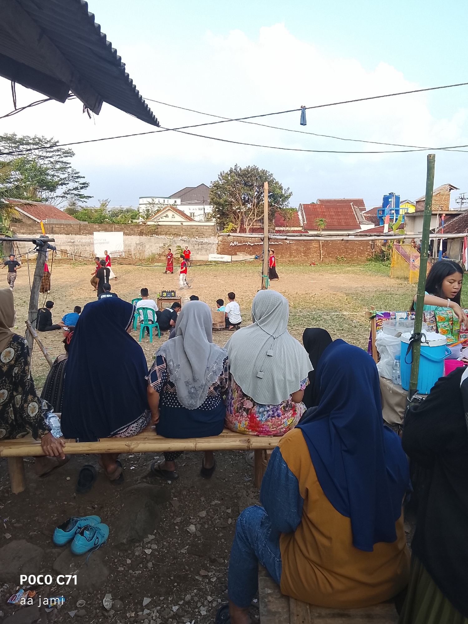 The image depicts an outdoor gathering, possibly a sporting event or community activity, taking place in what appears to be a rural or semi-urban area with some residential buildings in the background. Several people are seated on a bamboo bench in the foreground, facing away from the camera towards a dirt field where others are present. The atmosphere suggests a casual event, with some individuals standing and observing the activities on the field. The presence of a makeshift stall with drinks and snacks on the right side of the image indicates that refreshments are being sold, adding to the communal feel of the scene. The overall setting, with its simple infrastructure and modest surroundings, is consistent with a location in Tasikmalaya, Indonesia, where such local gatherings are common.