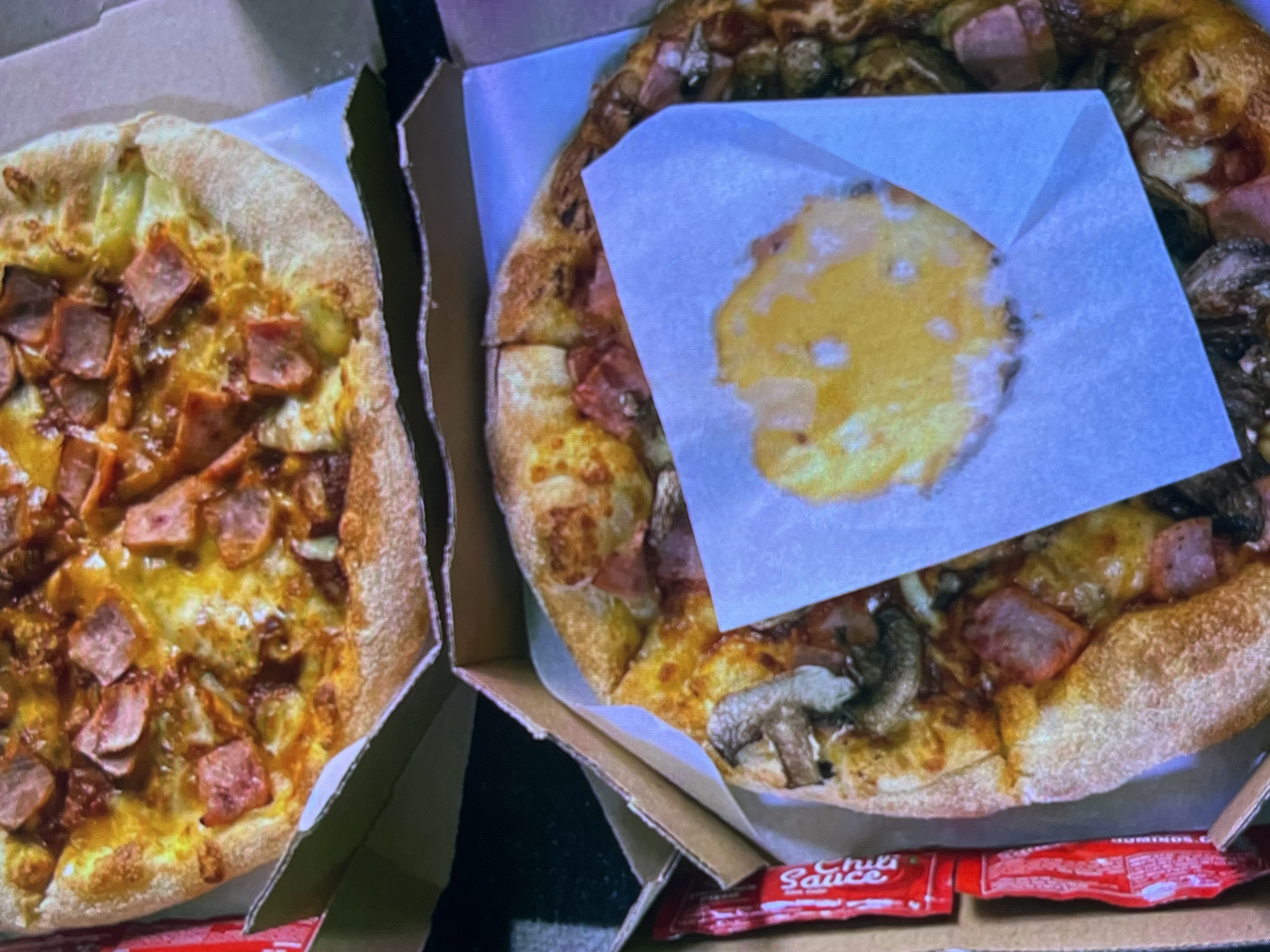The image shows two pizzas in open pizza boxes. The pizza on the left is mostly in focus and is topped with cheese, cubed ham, and some sort of sauce. The pizza on the right is slightly out of focus and has similar toppings of cheese and ham, as well as sliced mushrooms. A square piece of white paper is placed on top of the right pizza, with a piece of melted cheese on it.
The pizzas appear to be in a dimly lit indoor setting. The boxes are made of cardboard, and the background is dark, suggesting it might be nighttime or an indoor environment with low lighting. There are two red packets visible at the bottom of the image, one of which has the text "Chili Sauce" printed on it. The overall impression is of a casual meal being served or delivered. There are no people visible in the image. The pizzas look ready to eat.