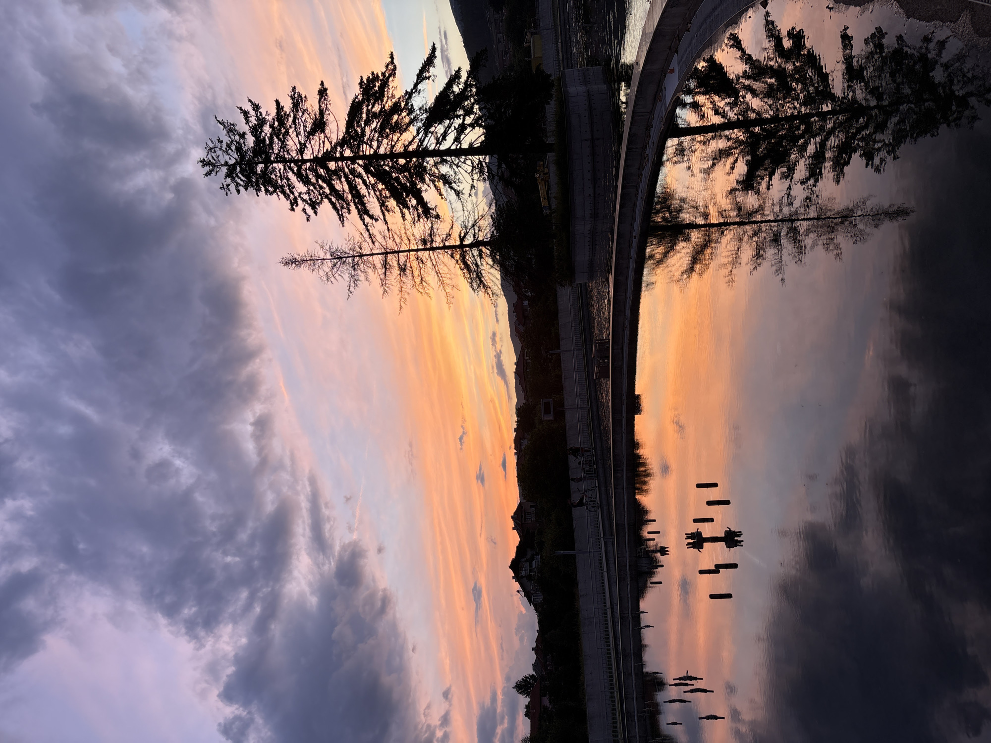 This photograph captures a serene sunset scene at the "Panagyurishte Water Mirror" in Panagyurishte, Bulgaria. The sky is a dramatic canvas of purples, pinks, and oranges, with scattered clouds tinged by the fading sunlight. Silhouetted against this vibrant sky are two tall pine trees situated on a small, elevated island within a large circular pool. The surface of the water perfectly reflects the colorful sky and the dark silhouettes of the trees and surrounding landscape, creating a symmetrical and tranquil composition. In the middle ground, a concrete walkway with railings runs along the edge of the pool, and several indistinct figures can be seen walking on it. The background reveals a residential area with houses and greenery nestled against rolling hills, all softened by the twilight. The water itself shows reflections of dark, rod-like structures emerging from its surface, adding another layer of visual interest to the scene.