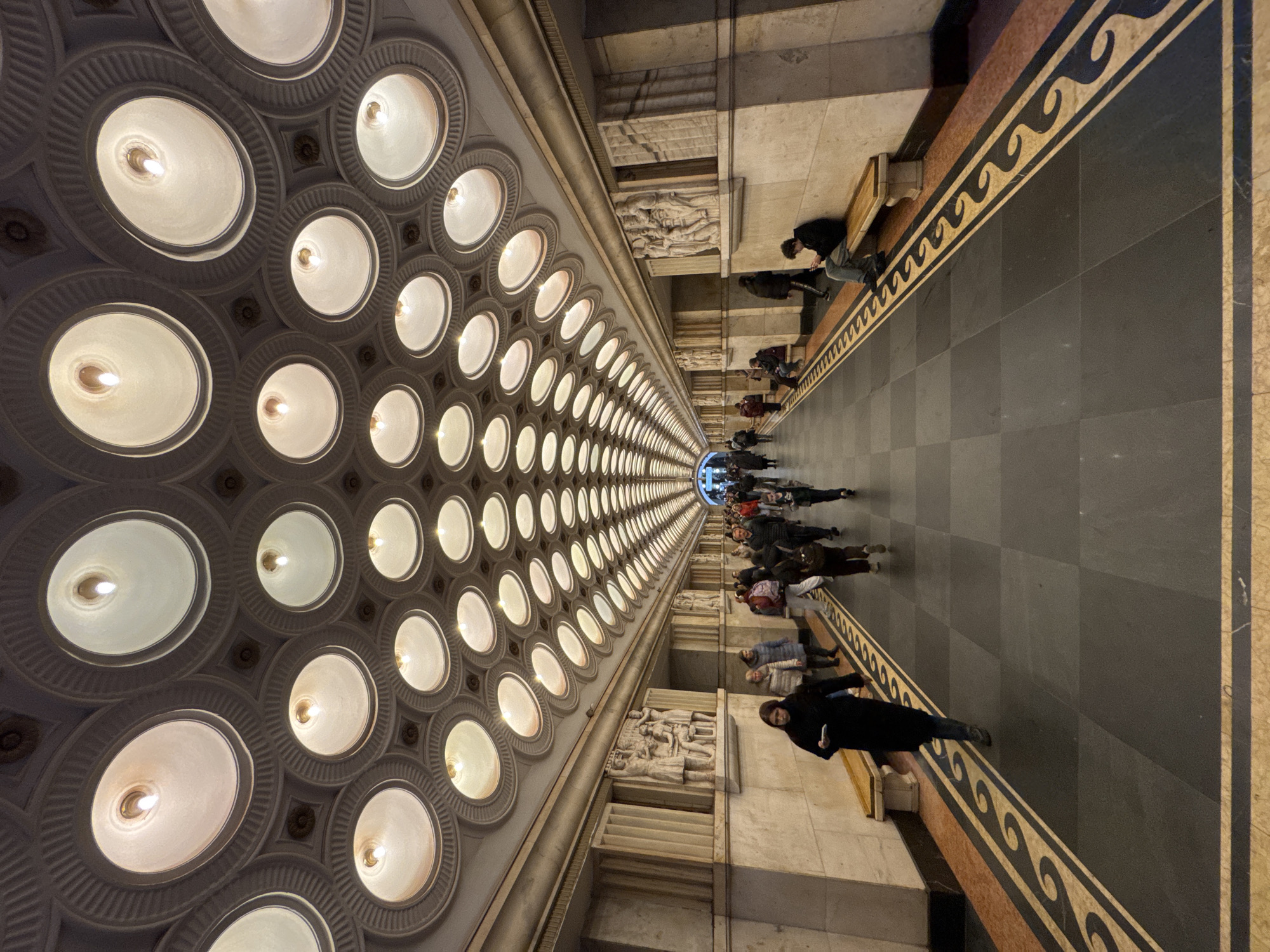 Here is a description of the image:
The image shows an interior space, likely a corridor or hall, with a distinctive ceiling featuring a grid of circular light fixtures. People are walking along the corridor. The walls are decorated with sculpted relief panels. The floor has a patterned design.
