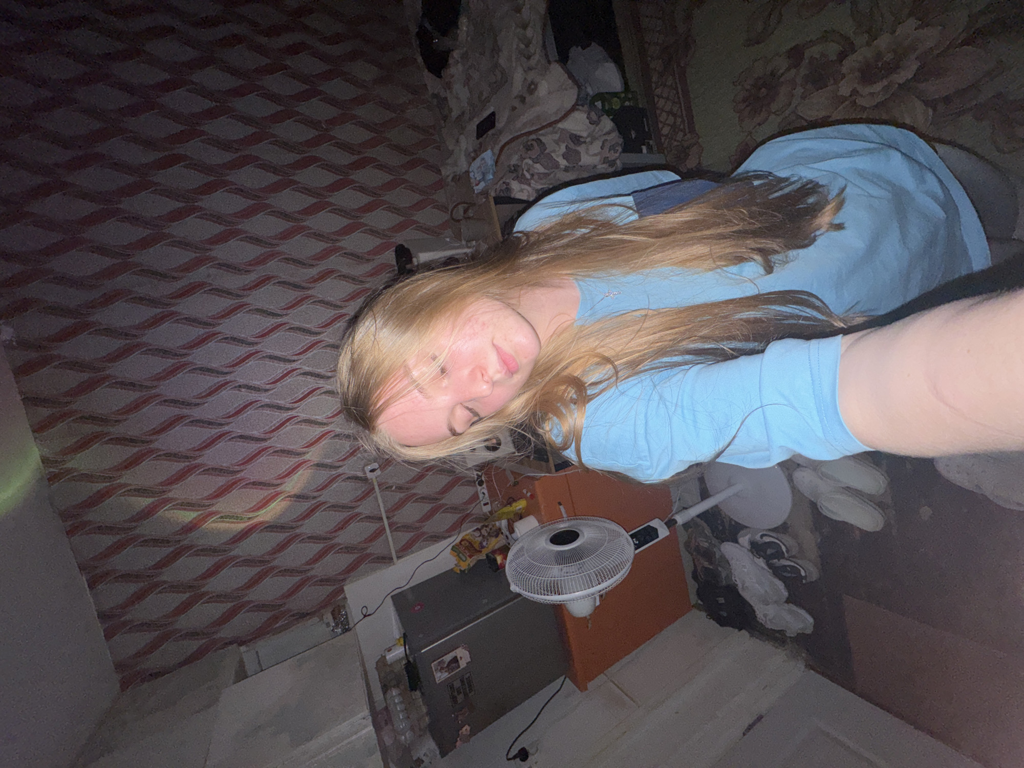 A young woman with long blonde hair lies on a bed in a room. She wears a light blue shirt. The room has patterned wallpaper and a patterned rug. A small fan sits on an orange and gray cabinet next to the bed. Shoes are visible on the floor near the cabinet. The photograph was likely taken in Perm, Russia, but this cannot be definitively confirmed from the image alone.