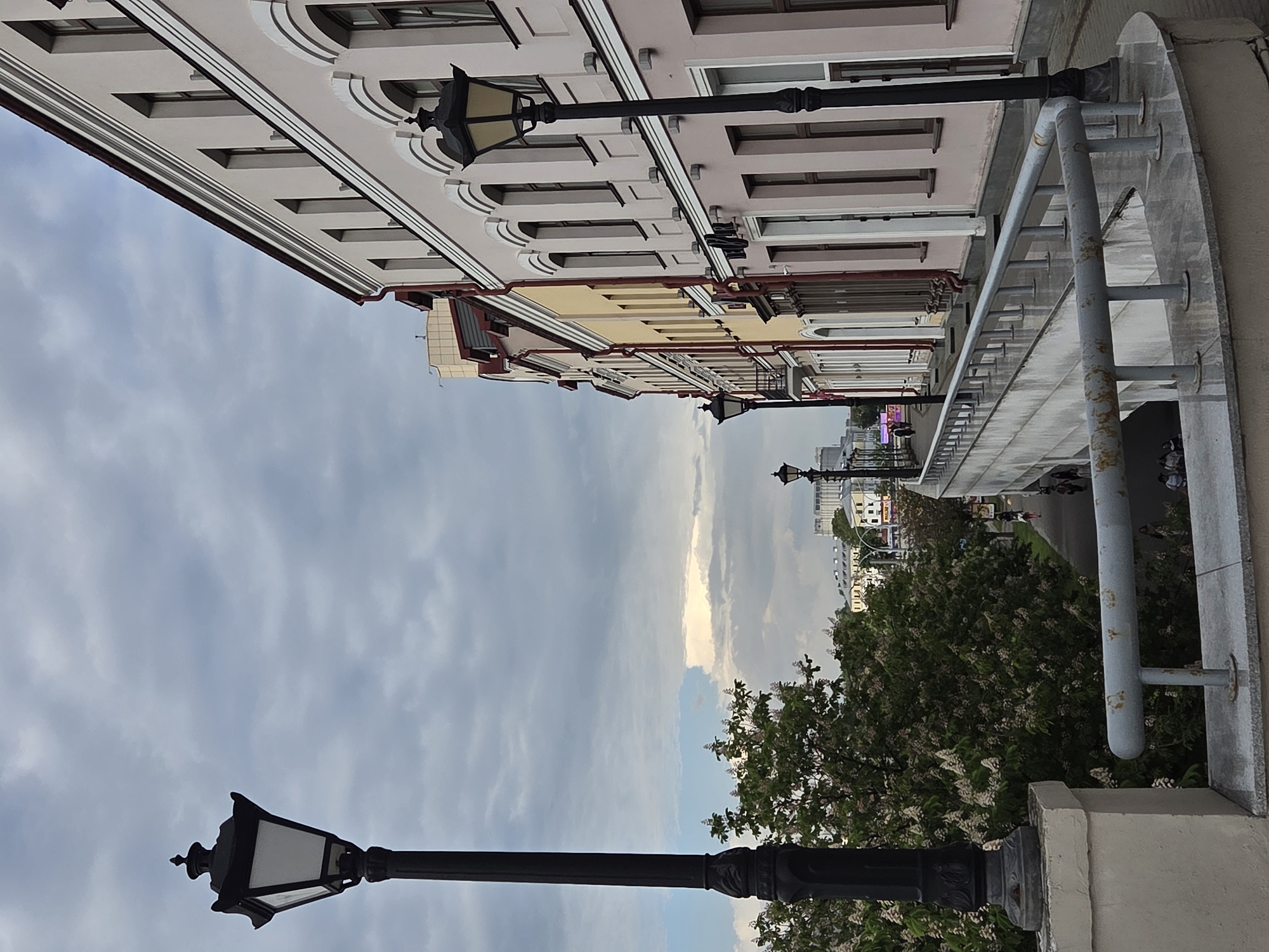 The scene depicts a streetscape in the city of Unknown, Unknown, viewed from an elevated position, potentially a balcony or bridge. The sky is overcast with grey and white clouds. Buildings line the street, appearing to be several stories tall, with architectural details like windows, cornices, and rooflines. A series of black lampposts, each with a classic design, are positioned along the street, adding to the vintage feel of the cityscape. A gray railing is visible in the foreground, suggesting the presence of a pedestrian walkway or platform. Below, a row of trees with white blossoms adds a natural element to the urban setting. Some people are walking on the sidewalk in the distance.