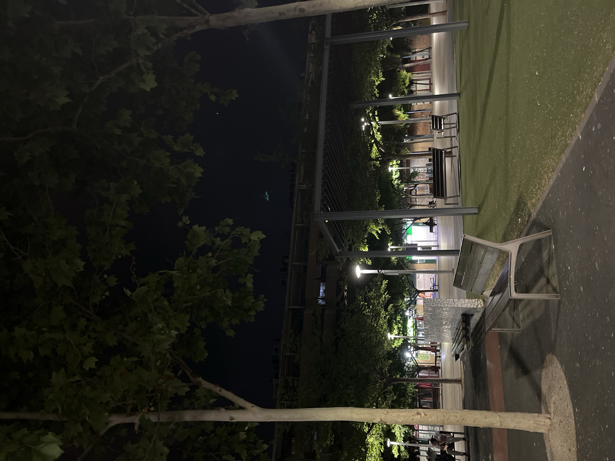 At night, a scene in Torrent, Spain reveals a partially illuminated outdoor space. Metal benches line a paved path alongside a patch of artificial turf, with trees bordering a dark body of water. Overhead, a metal pergola structure spans the area, and streetlights cast a glow on the surroundings. Pedestrians are visible in the distance near establishments with lit signs. The darkness of the night sky and the water contrasts with the lights of the city.