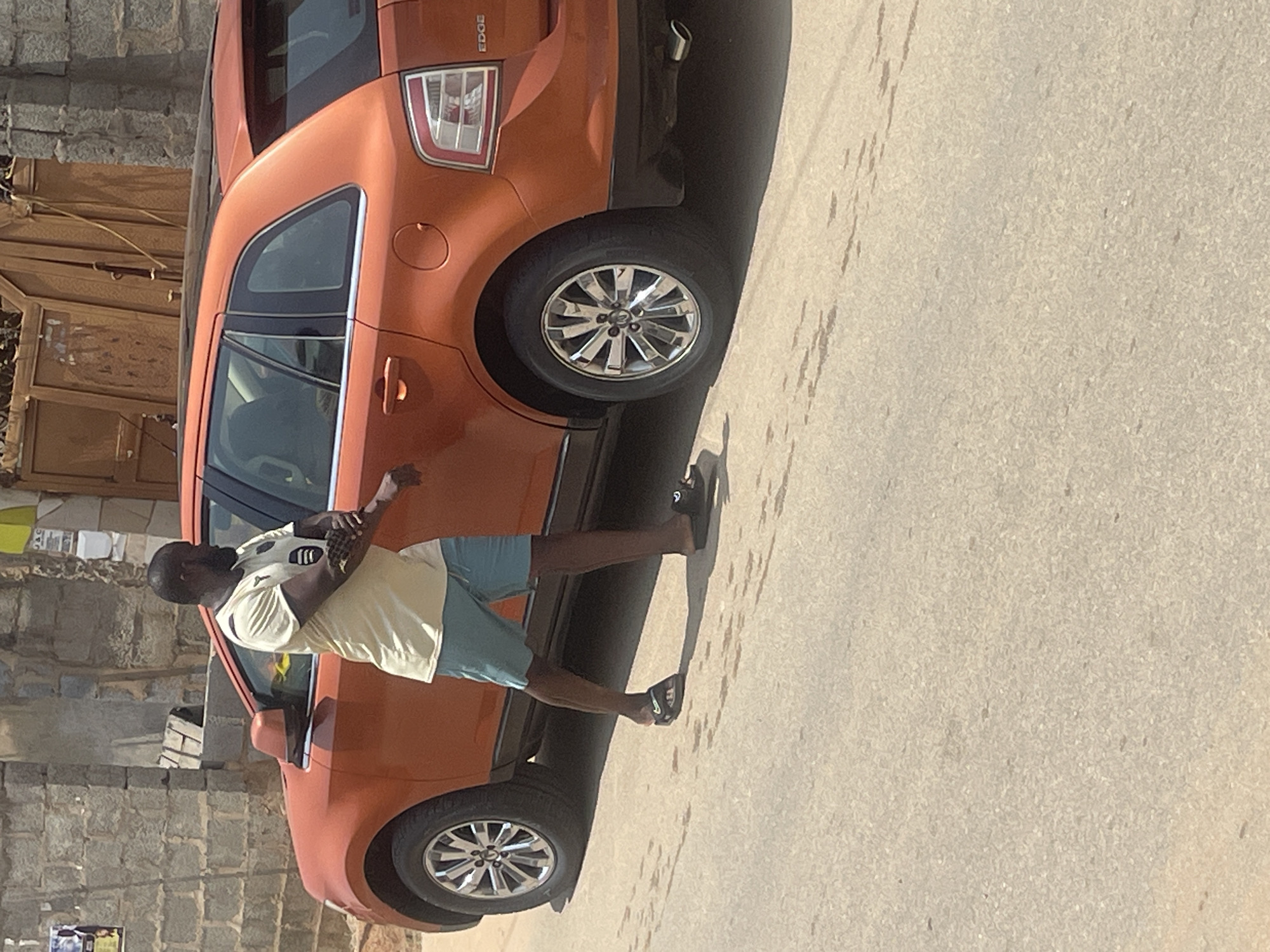 A man stands near an orange Ford Edge SUV. The car is parked on a paved surface, possibly a street or driveway. The man is wearing a light-colored t-shirt with dark stripes on the sleeves and light blue shorts. He is holding a white object with dark markings in his hands, which appears to be a ball. The man is looking towards the car.
Behind the car and to the left, there is a stone wall and a wooden structure that could be a gate or part of a building. The scene suggests an outdoor setting. The lighting indicates it is daytime, likely with clear weather. The text "EDGE" is visible on the rear of the car, indicating the model. The overall impression is of a casual moment, possibly before or after playing with the ball. The location context of Abuja, Nigeria, provides a geographical setting for this scene.