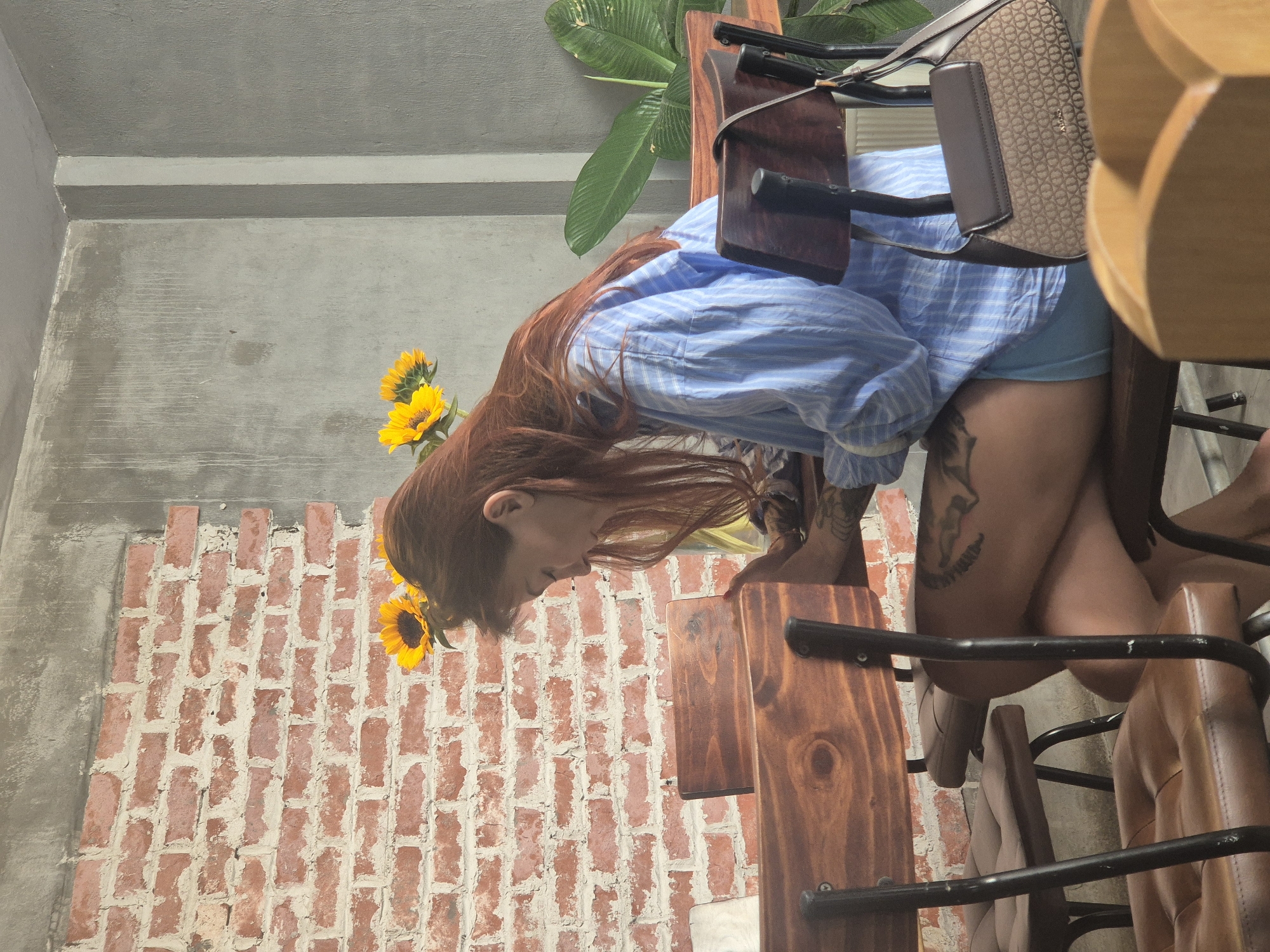 The image depicts a woman with long, auburn hair seated at a wooden table in a cafe. She is wearing a blue and white striped button-down shirt and light blue shorts, with a patterned handbag resting on the back of her chair. The cafe features exposed brick walls and a concrete accent wall. Two sunflowers are visible in a vase behind her. The scene appears to be in Đà Nẵng, Vietnam.