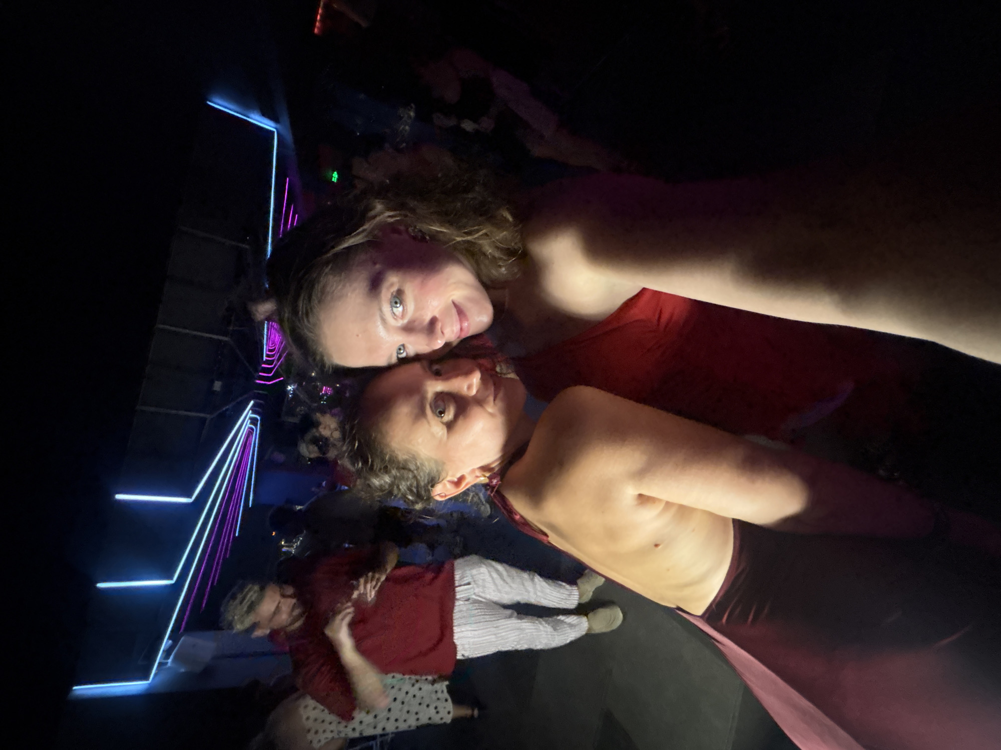 The image shows two women in a dimly lit nightclub. The woman on the left is wearing a red dress, and the woman on the right is wearing a red one-shouldered top. Both women are smiling and looking at the camera. There are other people in the background, some of whom are dancing. The nightclub has neon lights in blue and pink. The image does not provide any specific details about the city of Đà Nẵng, Vietnam.