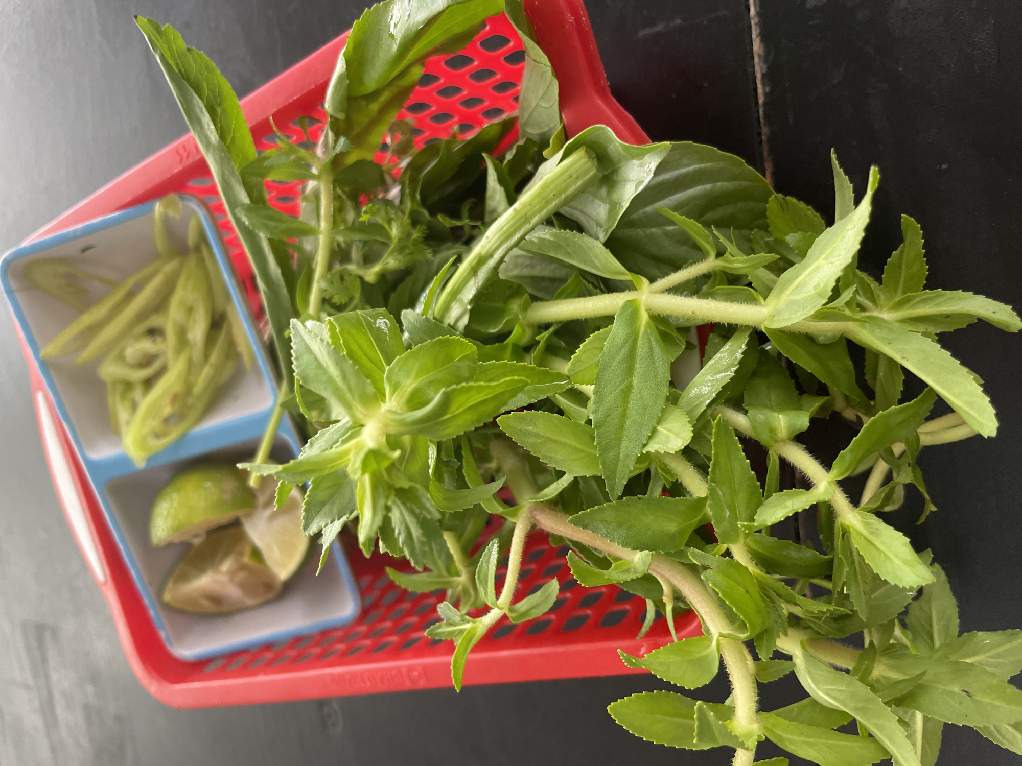 A close-up, high-angle shot shows a red plastic basket containing various herbs and garnishes. In the upper left compartment of the basket, there are two wedges of lime. In the compartment to its right, there are slices of green chili pepper. The majority of the basket is filled with fresh green herbs, primarily basil and what appears to be Vietnamese mint or perilla. The herbs are vibrant green and have a fresh, slightly dewy appearance. The basket is placed on a dark, textured surface, likely a wooden table. The background is dark and out of focus, suggesting an indoor setting, possibly a restaurant in Phu Thuy Ward, Vietnam. There are no people or animals visible in the image.