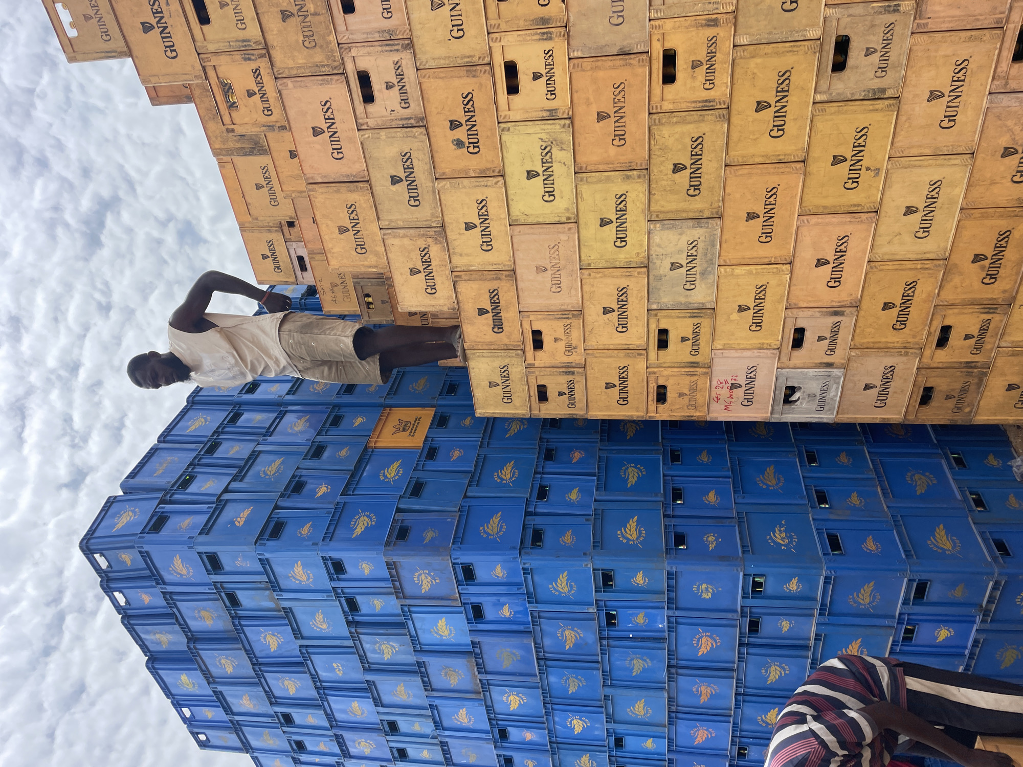 In Bukuru, Nigeria, the image depicts a stack of blue "Premium Brew" beer crates forming a wall or base, topped by a wall of tan "Guinness" beer crates. A dark-skinned man in a light-colored shirt and shorts stands atop the "Guinness" crates, appearing to survey the scene. Another person, partially visible in the lower right corner, wears a striped shirt. The sky with clouds is visible above.
