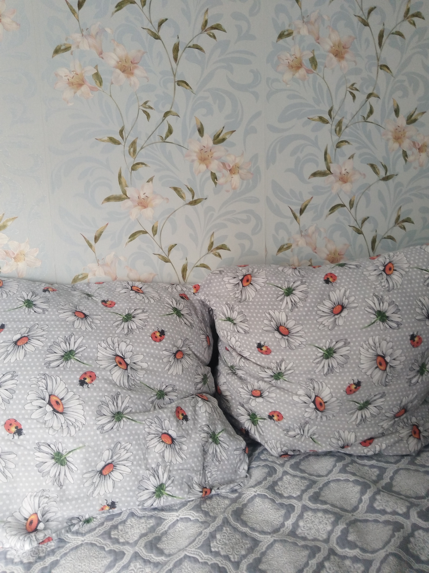 Two pillows with a daisy and ladybug pattern are resting against a wall with floral wallpaper. The wallpaper features light blue swirls and pink and white flowers. The pillows are a light gray color with a repeating pattern of white daisies with orange centers and red ladybugs. The background wallpaper has a subtle floral pattern in shades of light blue and white, with hints of pink and green from the flowers and leaves. The image is a close-up shot, focusing on the textures and patterns of the bedding and wallpaper. No people are visible in the image.